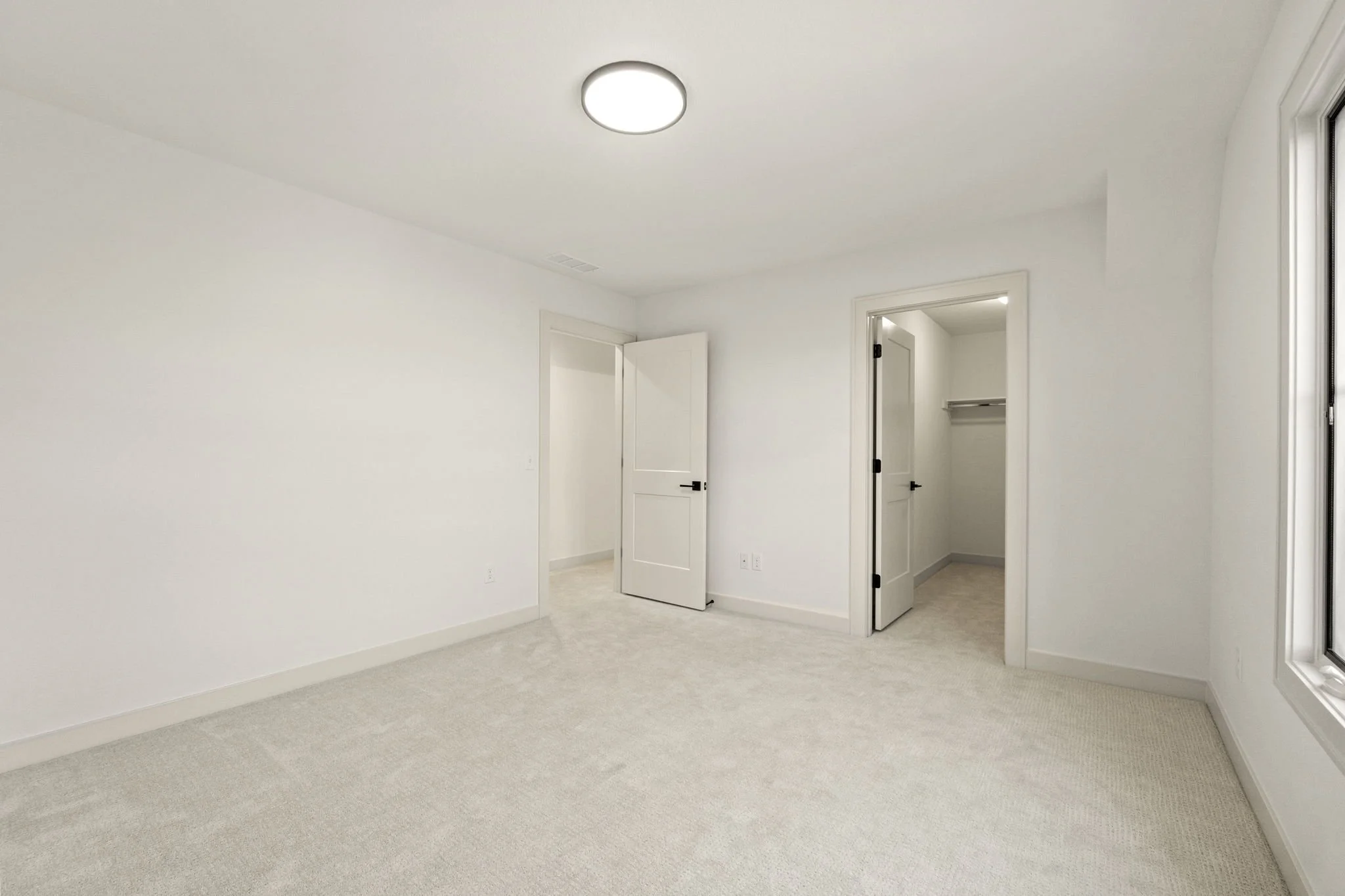Empty bedroom with white walls, beige carpet, ceiling light fixture, and two open doors leading to closet and hallway.