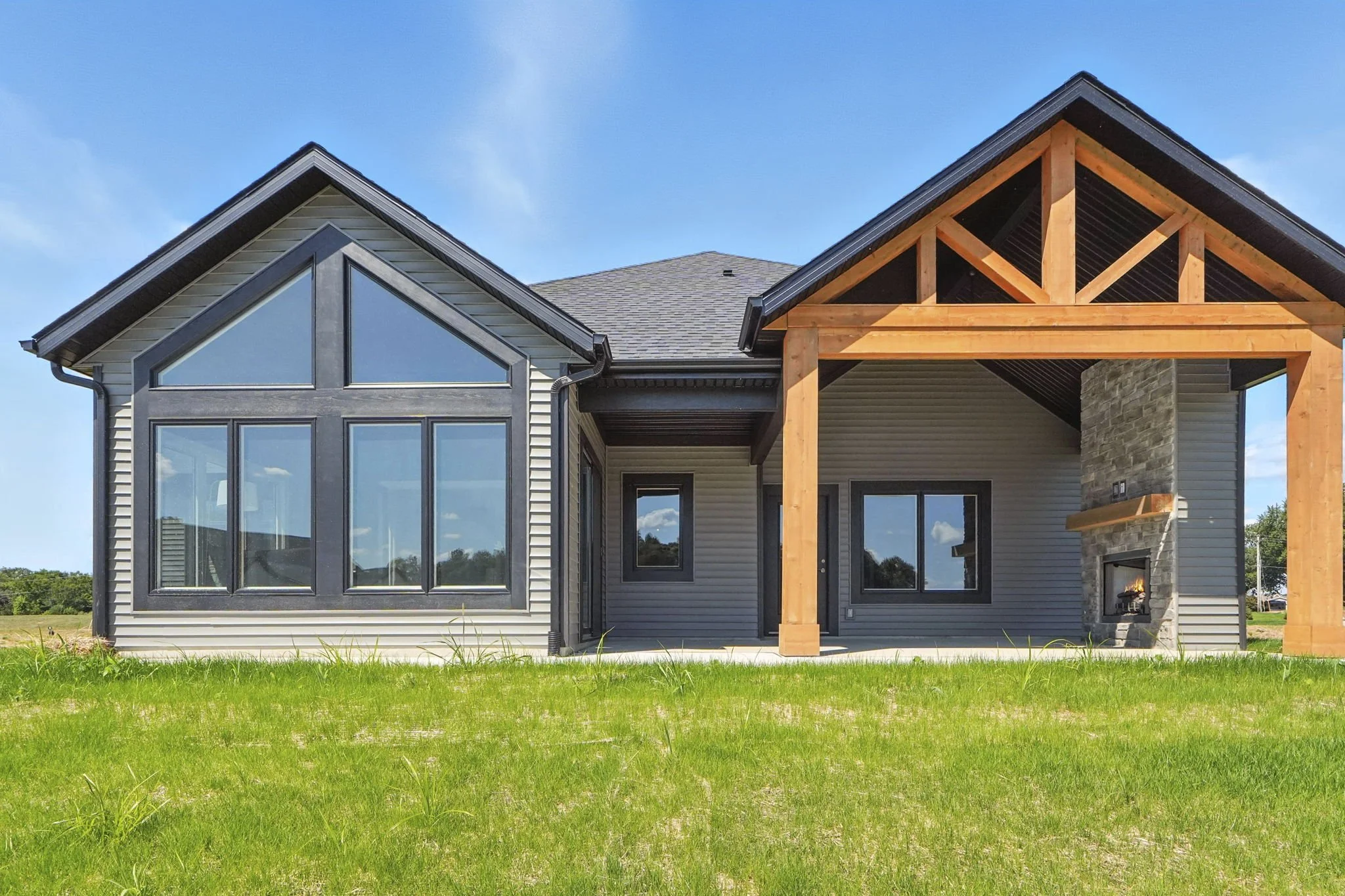 Modern house with large windows, vinyl siding, and a stone fireplace, situated on a grassy lawn with a blue sky background.