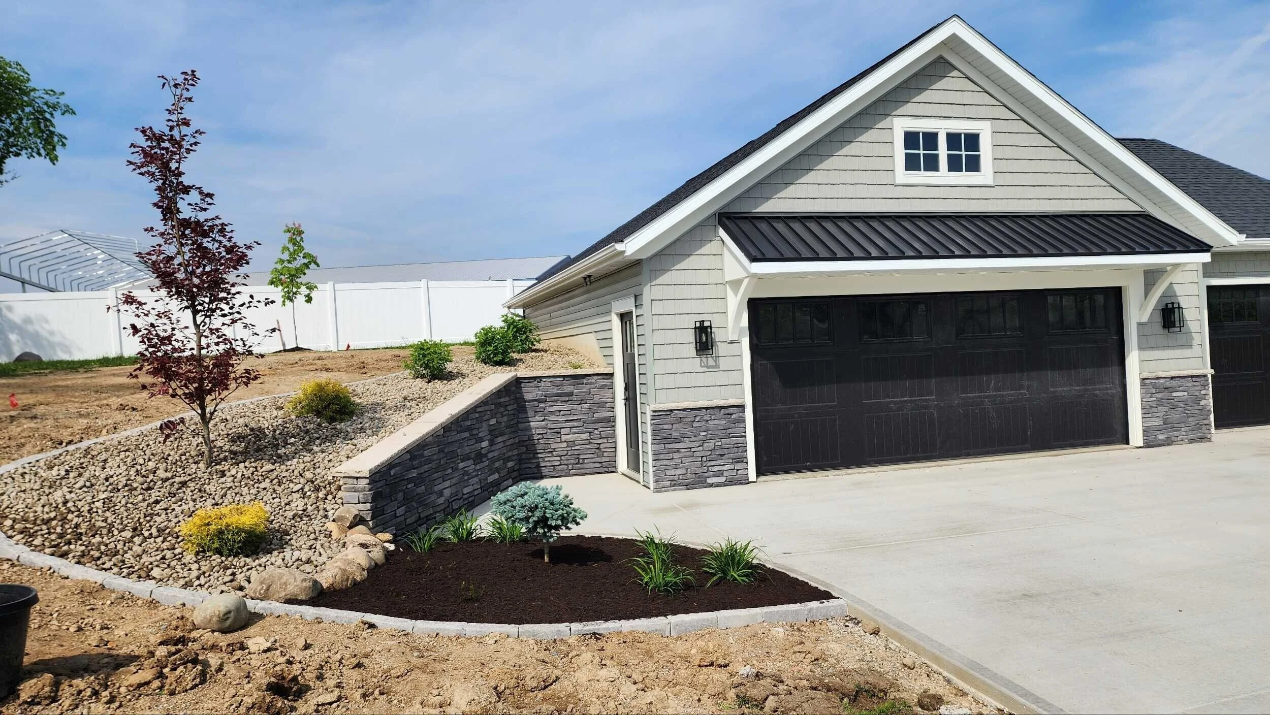 Newly built house with a black garage door, gray siding, and a landscaped front yard featuring small trees, shrubs, and gravel mulch under a blue sky.