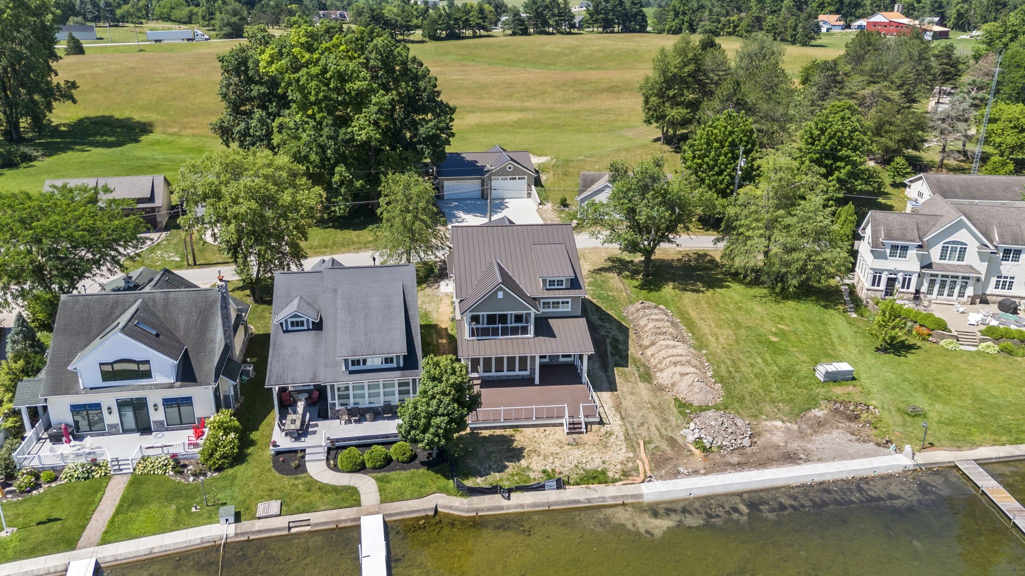 Aerial view of a residential neighborhood along a waterfront, featuring several large houses with well-maintained lawns and trees, and a construction site next to one house.