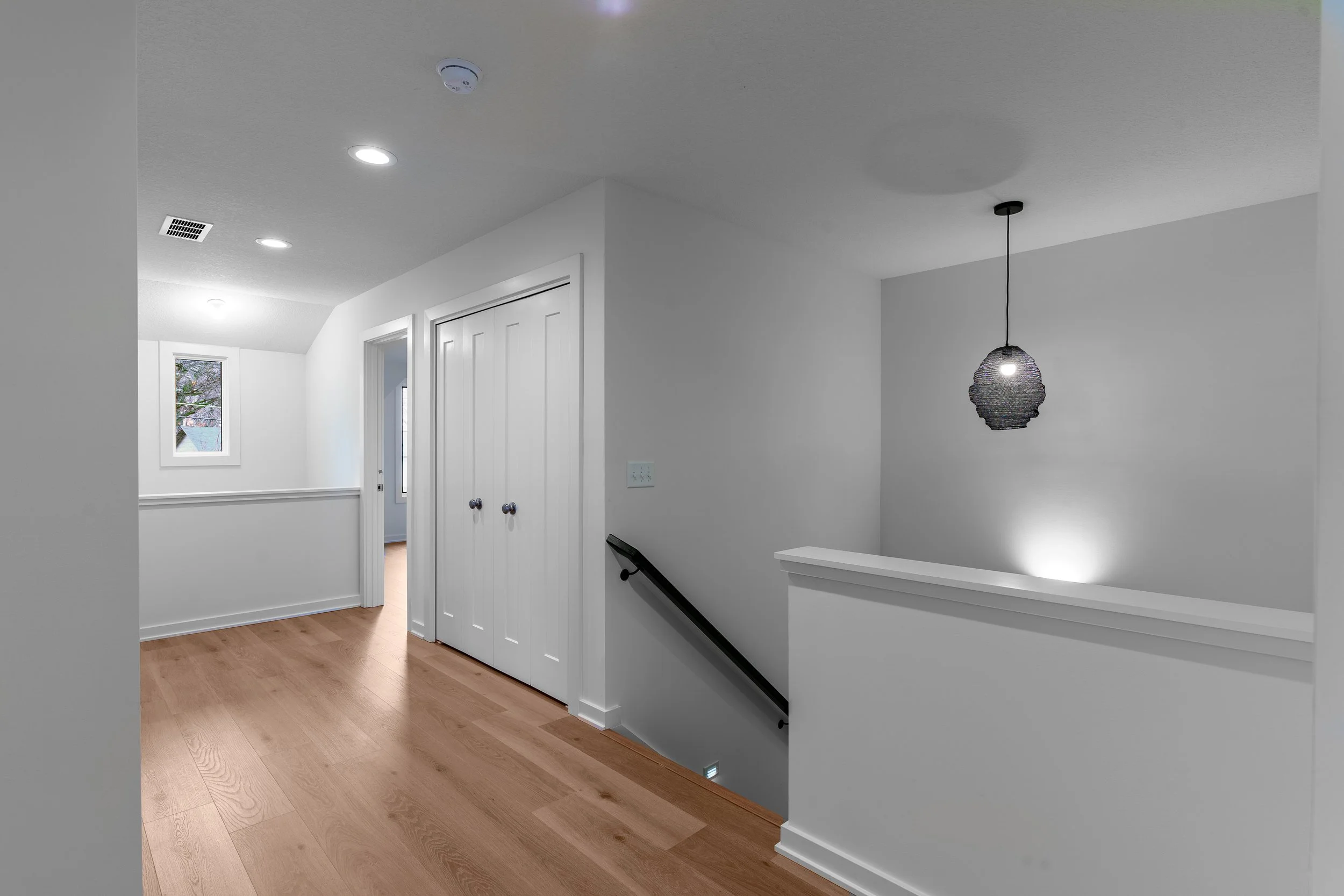 Interior view of a modern, minimalistic hallway with white walls, light wood flooring, a small window, ceiling lights, a staircase with a black handrail, and a pendant light fixture.