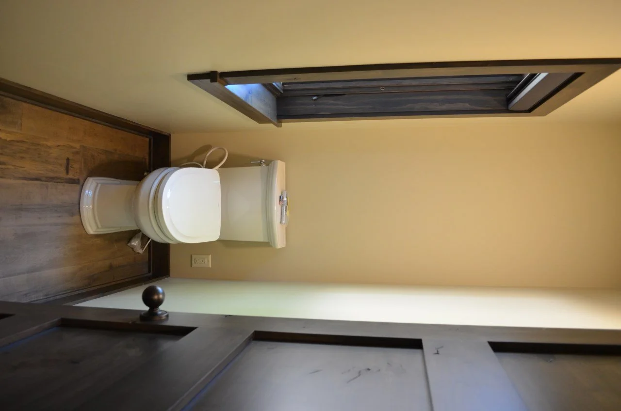 A small bathroom with a white toilet and a window above it, wooden flooring, and beige walls.