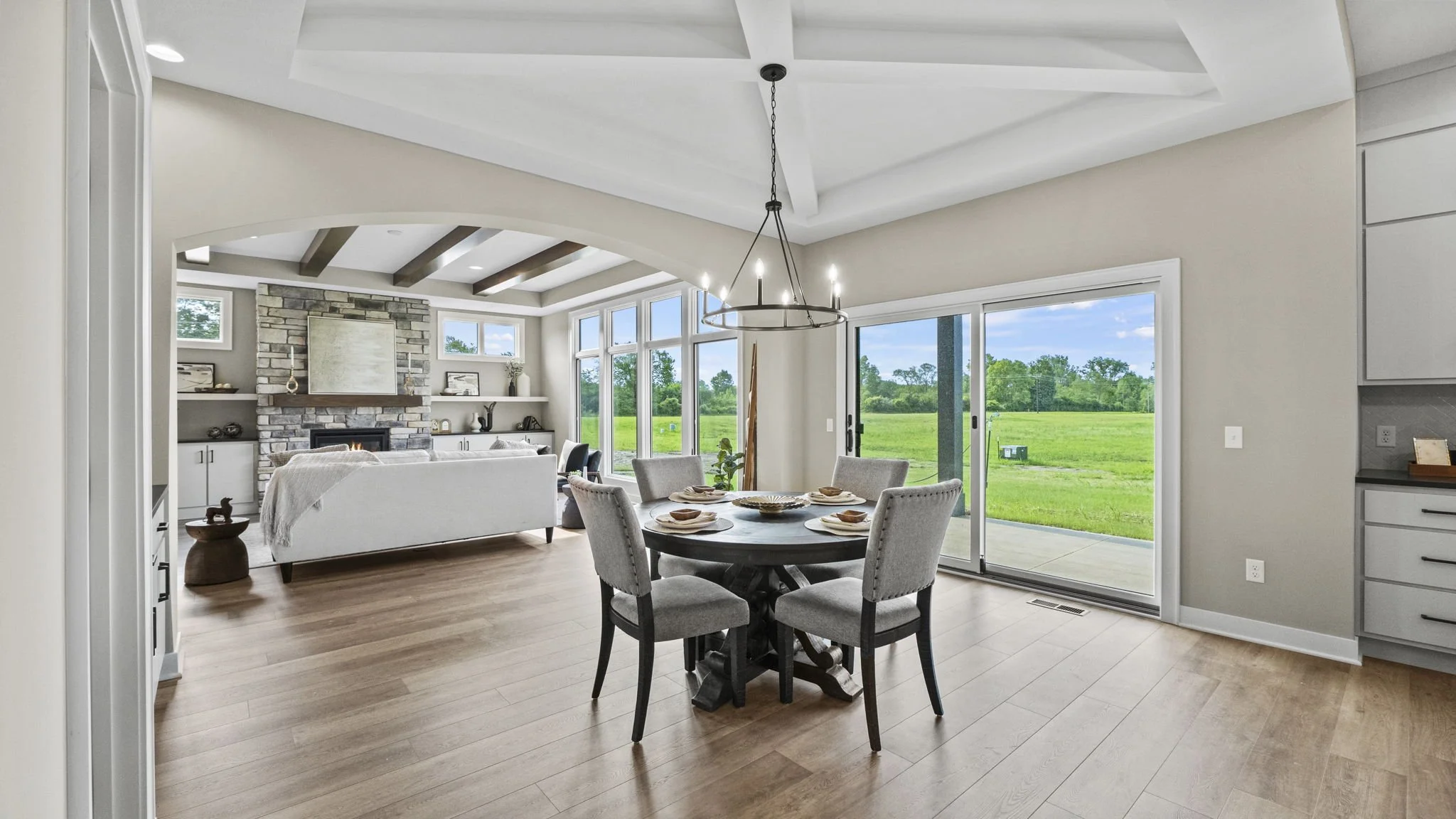Open living and dining area with large sliding glass doors opening to a grassy backyard, a round dining table with six chairs, and a stone fireplace with built-in shelves in the background.