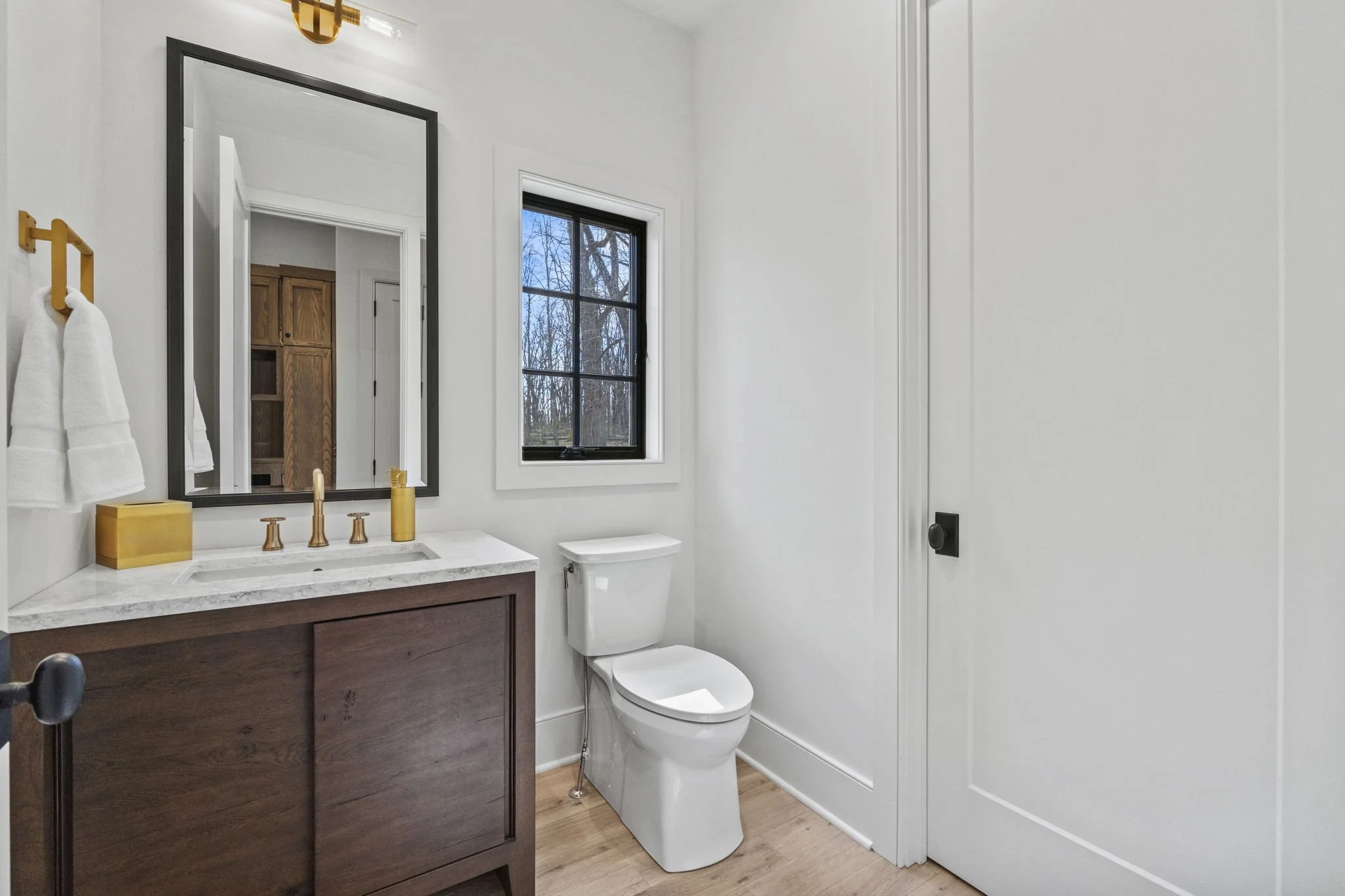 A modern bathroom with a white toilet, a wooden vanity with a marble countertop, a large mirror, a window showing trees outside, and white walls with simple decor.