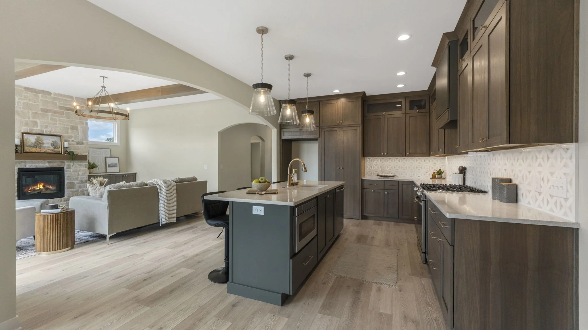 Open-concept kitchen with dark wood cabinets, white countertops, an island with a sink, three pendant lights, and a seamless transition to a living room with a stone fireplace and seating area.