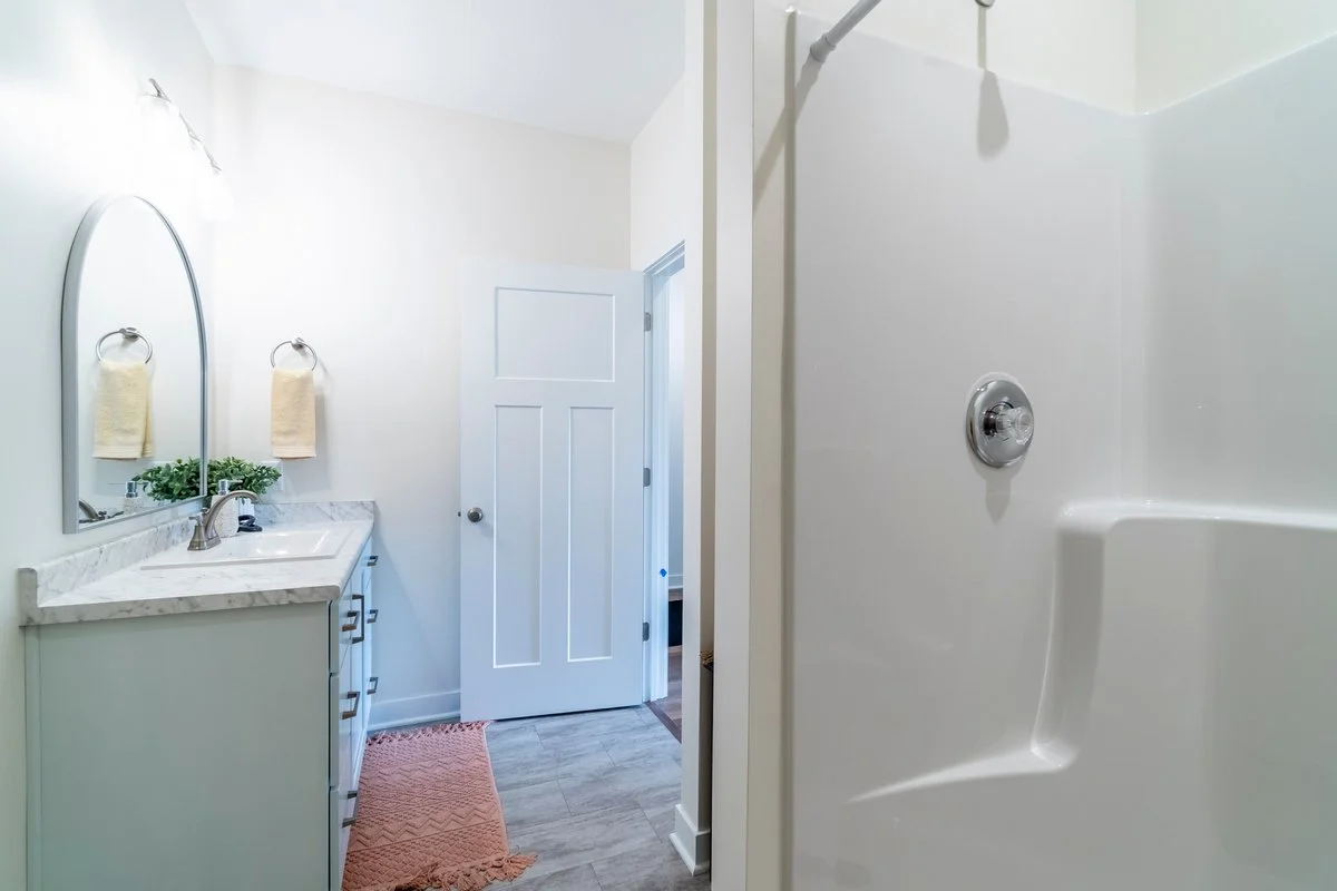 A bathroom with a white vanity, a mirror, towels, a plant, a door, and a shower stall.
