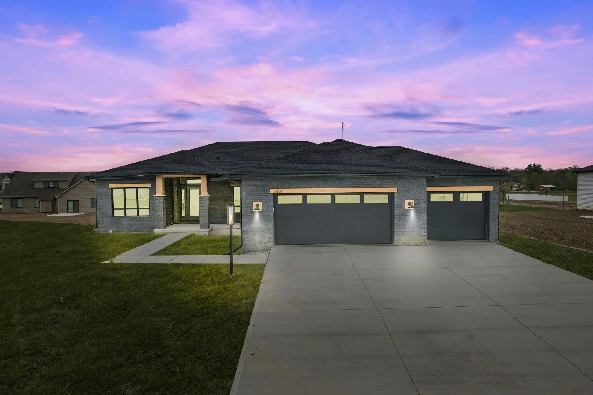 A modern single-story house with a dark gray brick exterior, large windows, and an attached three-car garage. The house is set against a colorful sunset sky with pink and purple clouds, and has a concrete driveway and a small front lawn.