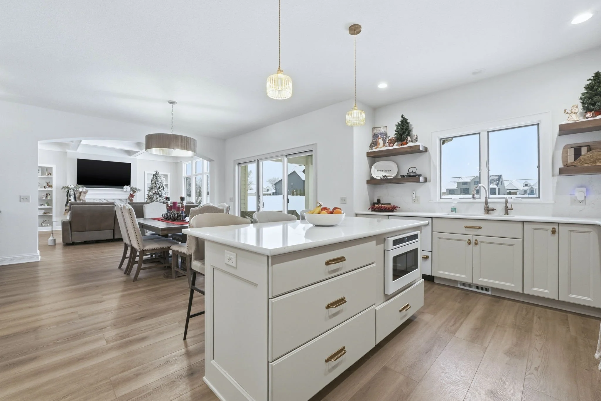 Bright open-concept kitchen with white cabinets and island, displaying a fruit bowl, with dining area and living room in background, decorated for Christmas.