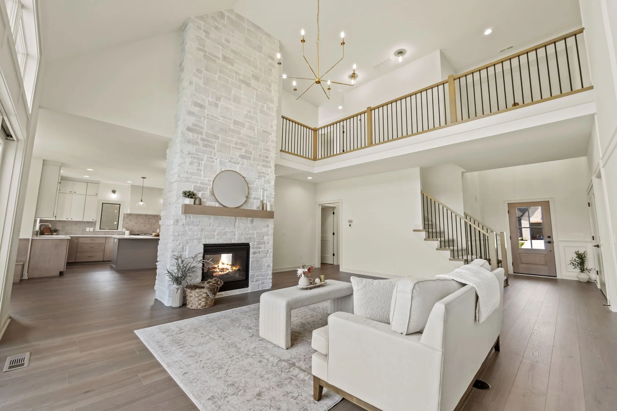 Open-concept living room with white walls, wood flooring, a stone fireplace, modern chandelier, beige sofa, coffee table, and staircase leading to a second floor with wooden railing.