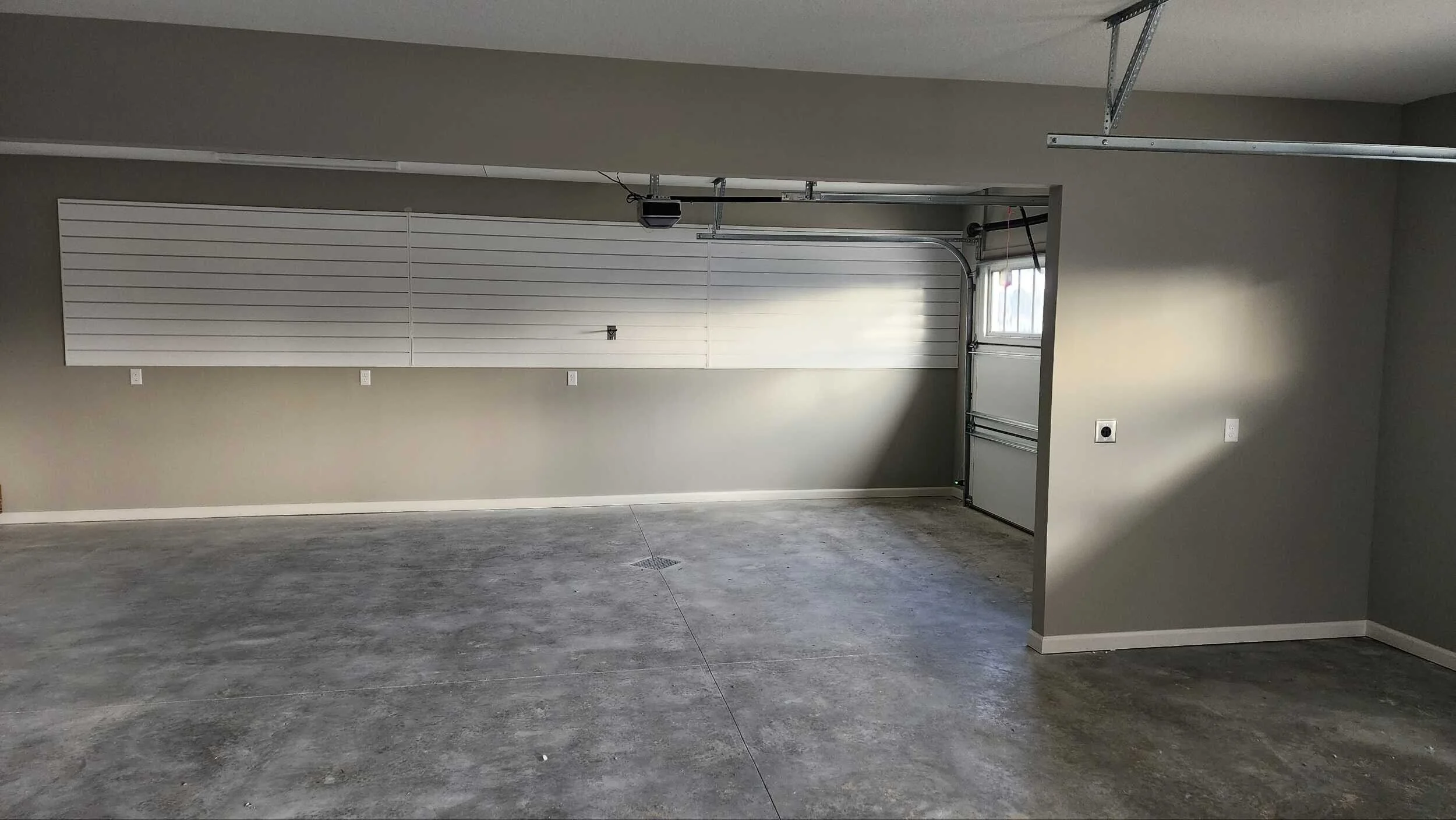 Empty garage interior with concrete floor, gray walls, a partially open garage door with wall-mounted shelving and electrical outlets.