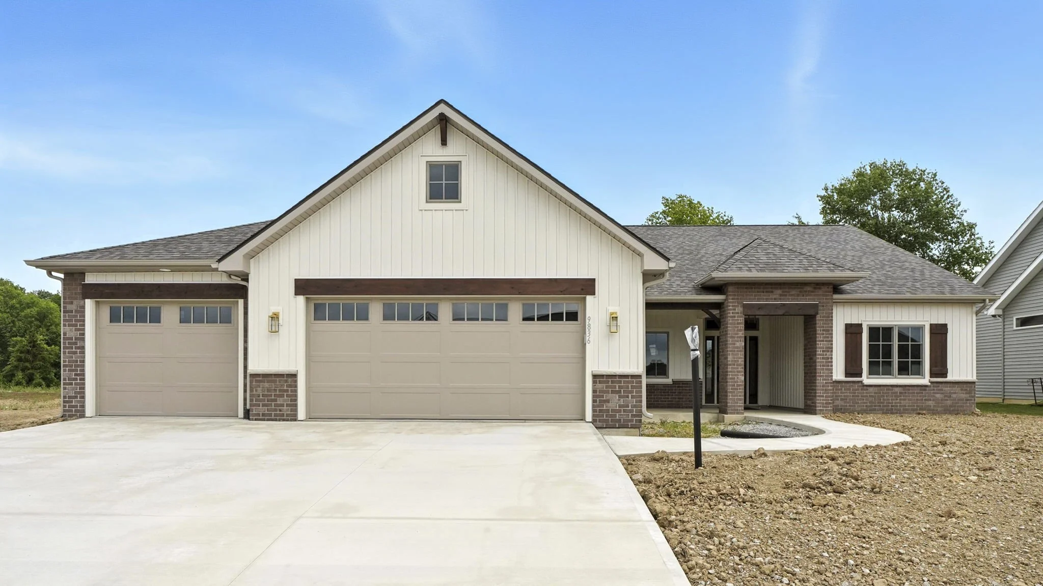 Newly constructed modern house with a three-car garage and a mixture of white vertical siding and brick exterior, with a concrete driveway, and some construction site dirt in the yard.