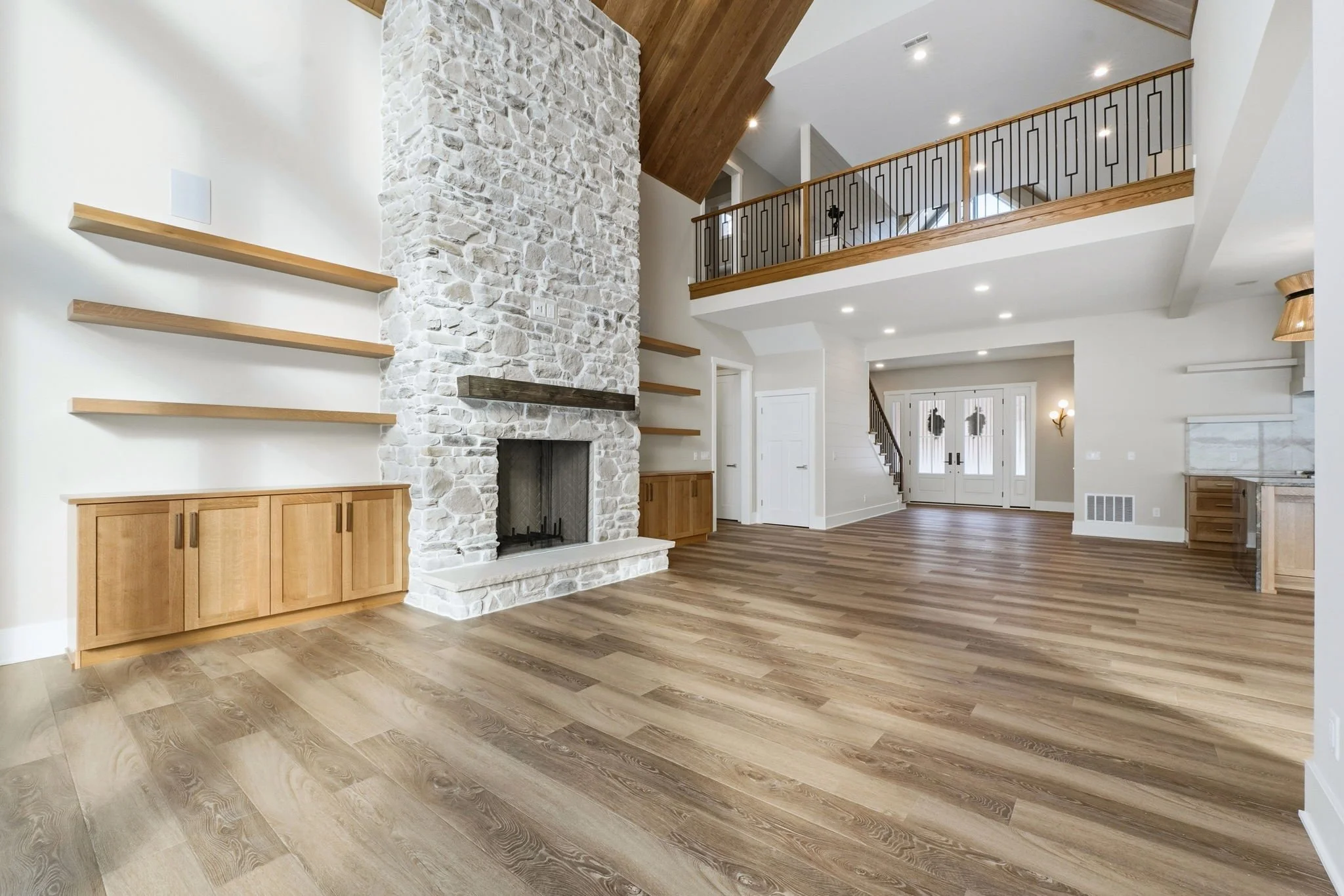 Empty living room with a white stone fireplace, light wood flooring, built-in wood cabinets, and an upstairs balcony with metal railing.