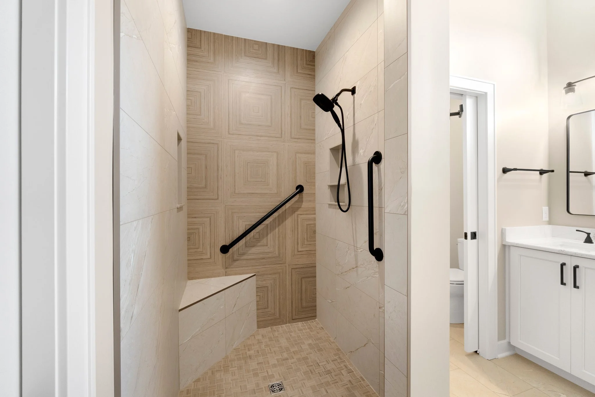 Modern bathroom with a walk-in shower featuring a beige tiled wall with geometric patterns and black grab bars, adjacent to a white vanity area with a mirror and a doorway to a toilet.