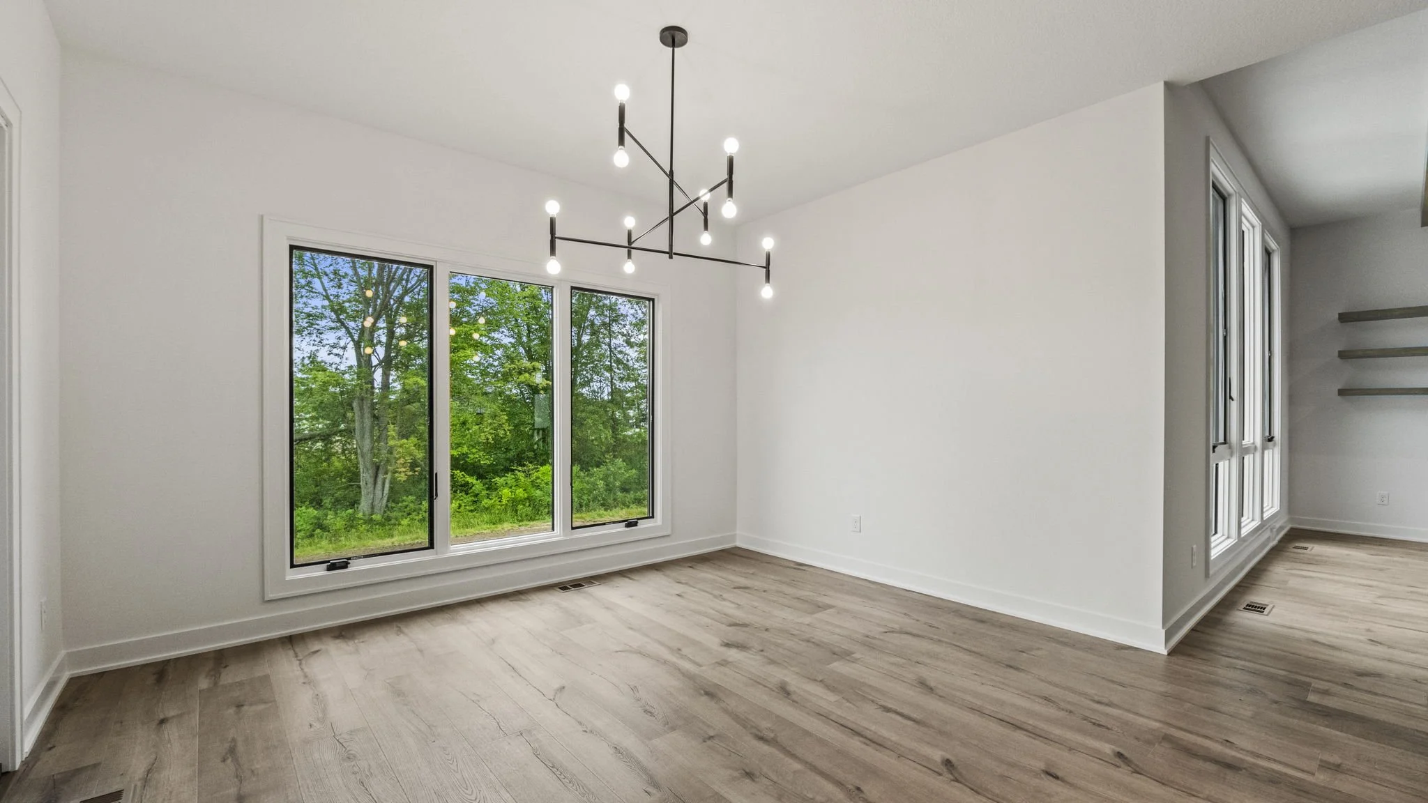 Empty modern living room with large window looking out to green trees, white walls, light hardwood flooring, and a contemporary chandelier with exposed bulbs hanging from the ceiling.