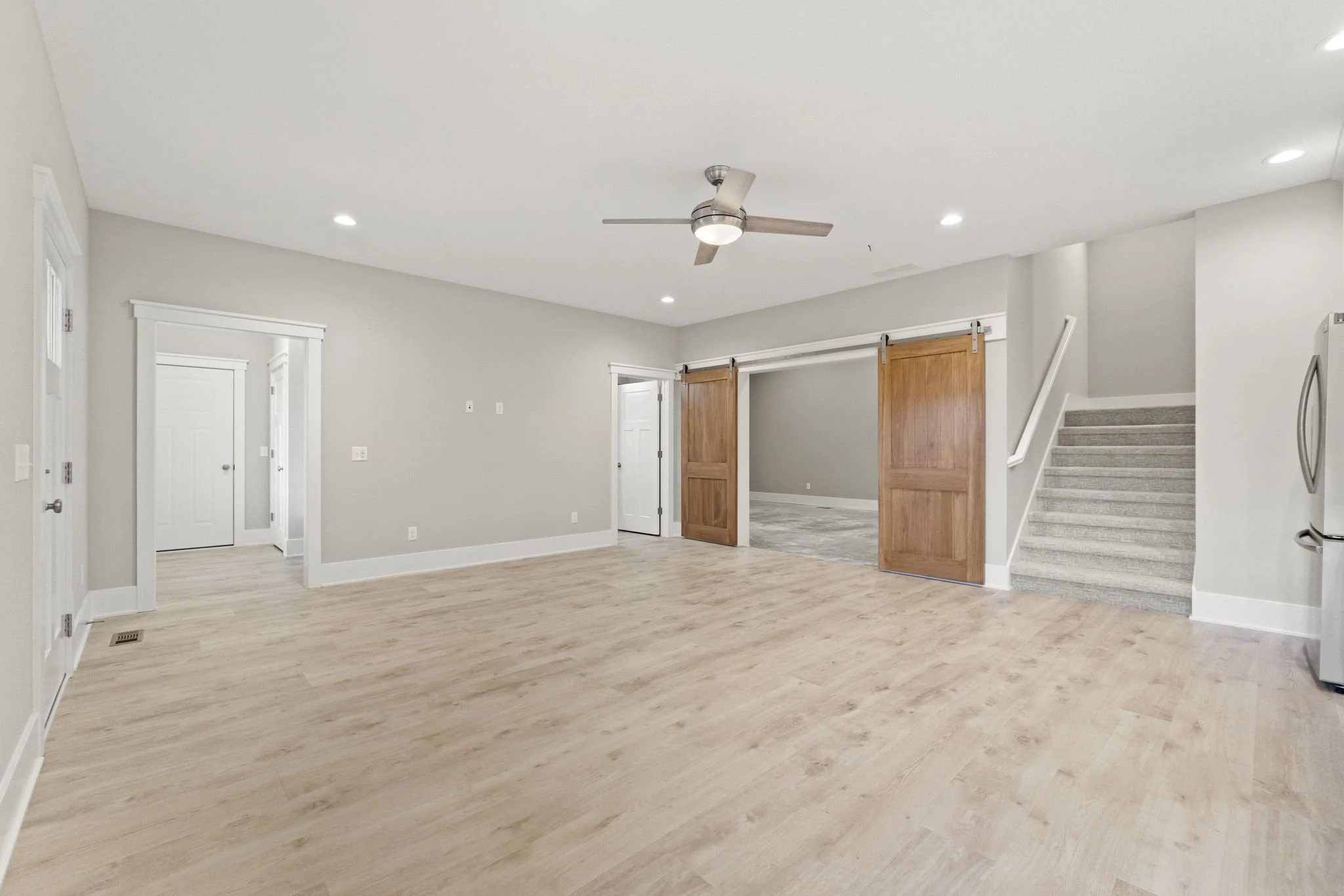 Empty living room with light-colored hardwood floors, white walls, a ceiling fan, and recessed lighting. There are sliding barn doors leading to another room, a staircase with carpeted steps, and a partial view of a kitchen refrigerator.