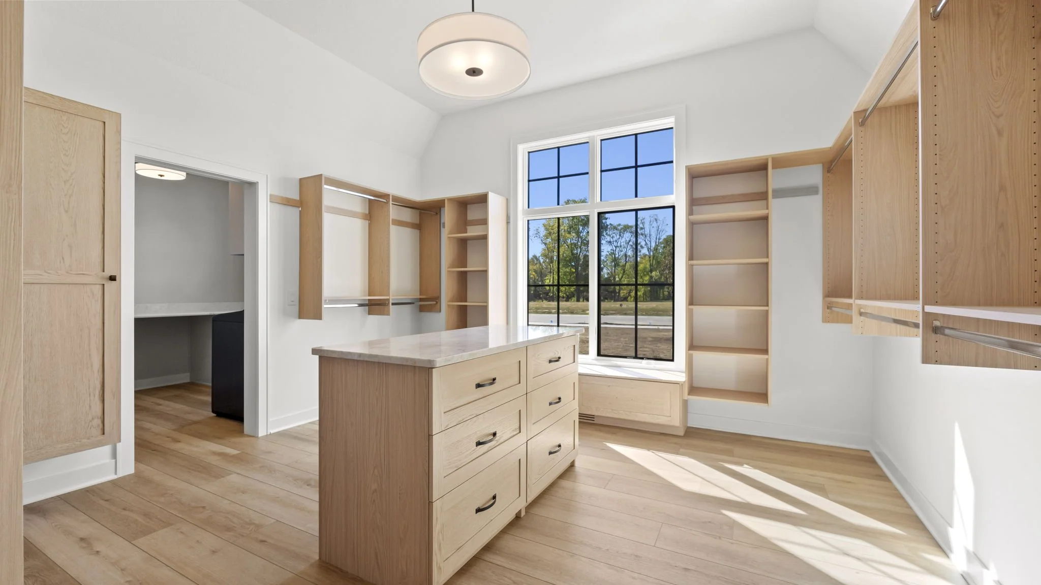 Empty walk-in closet with wooden shelves and drawers, large window with steel grid, hardwood floor, white walls, and ceiling light fixture.