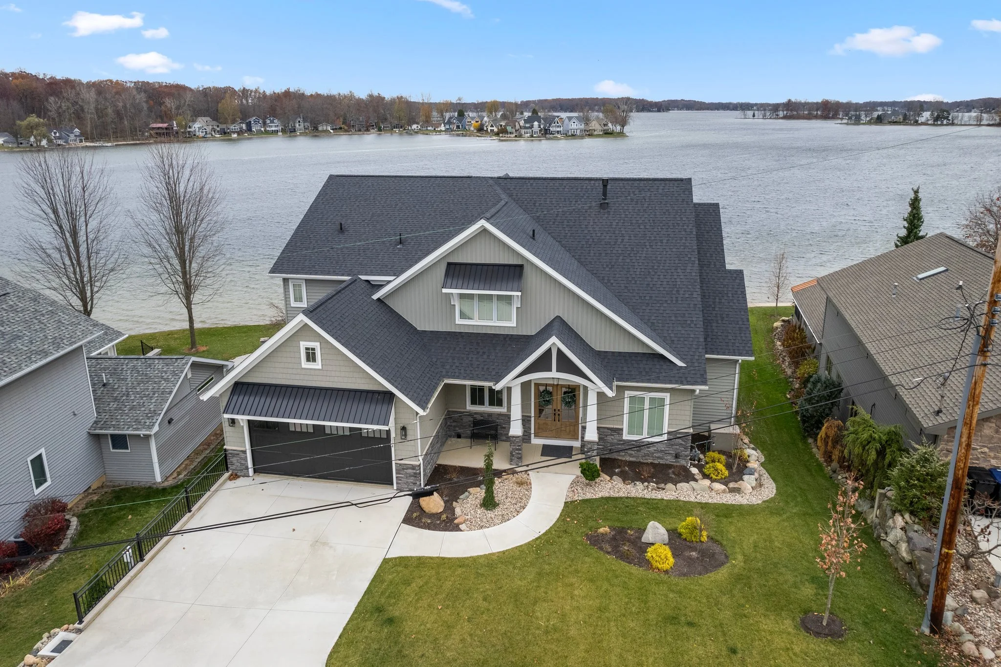 A modern house with gray siding and black roof, situated next to a lake, with a driveway and landscaped front yard.