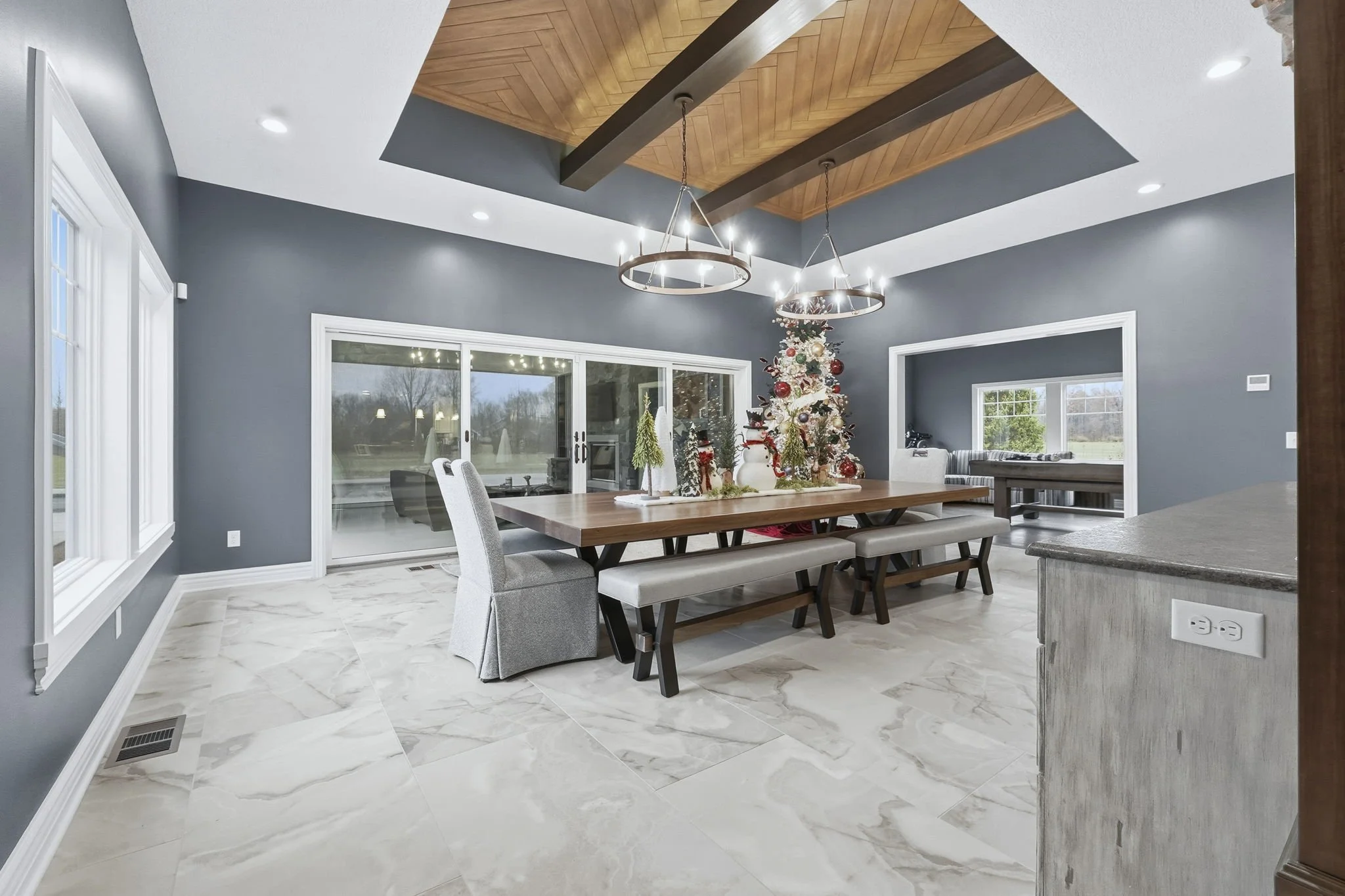 A spacious dining room with a modern design, featuring a wooden dining table decorated with a Christmas centerpiece including a Christmas tree, snowman figure, and other holiday decorations. The room has large windows and sliding glass doors, gray wa