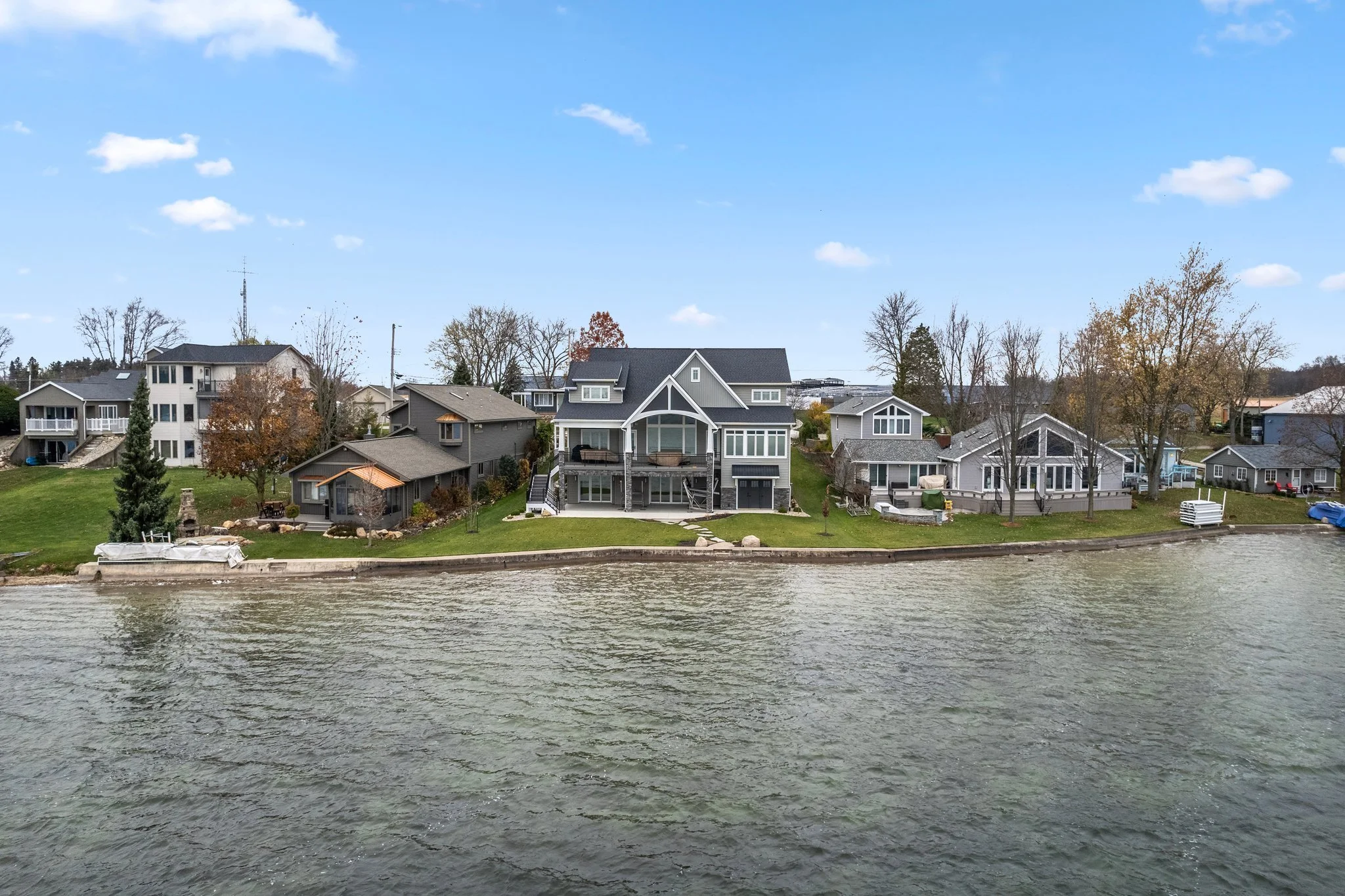 Residential houses along a lake with a grassy yard, some trees, and a blue sky with clouds.