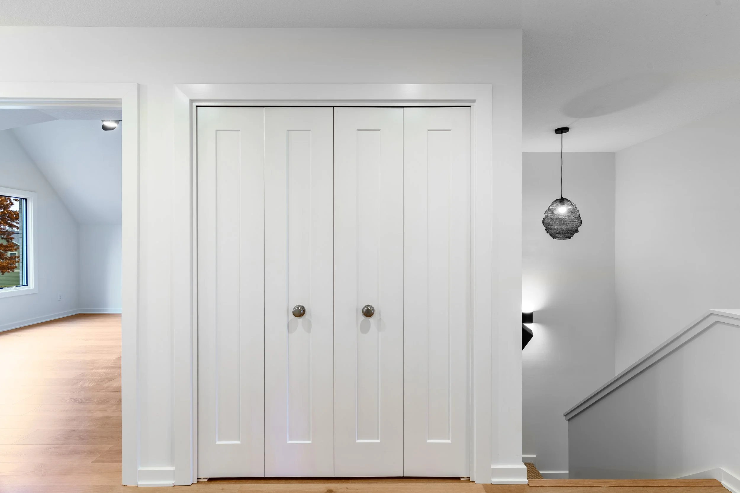 White double closet doors with silver knobs in a modern interior, partially showing a staircase with a black wall-mounted light fixture and a hanging black pendant lamp.