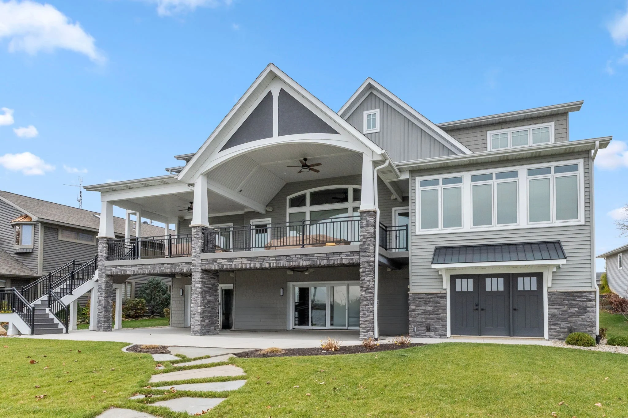 Large modern house with gray siding, stone accents, two upper balconies, a garage, and a well-maintained lawn.