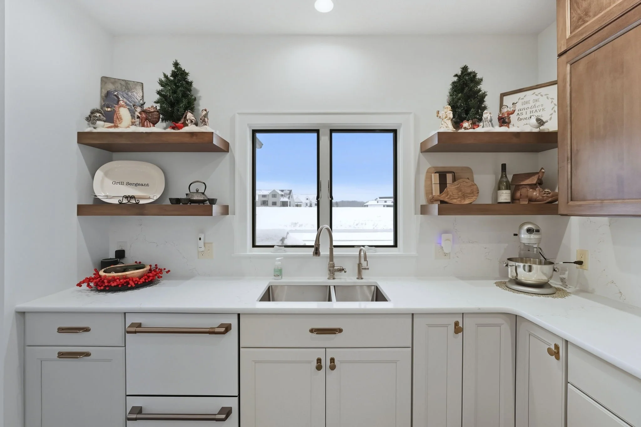 Kitchen with white cabinets, dual sink, window showing snow outside, and decorative Christmas shelves with figurines and greenery.