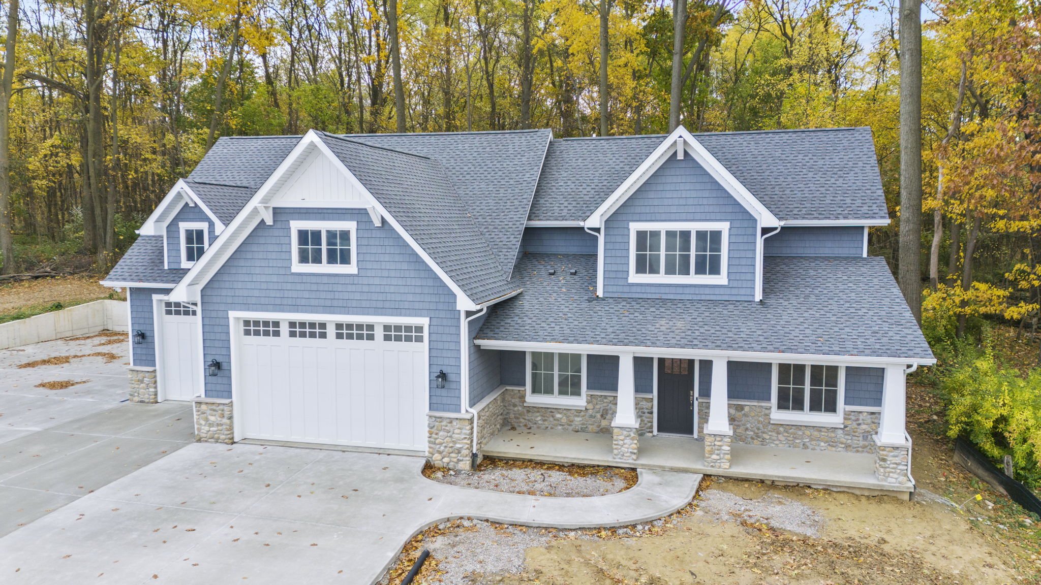 A newly constructed two-story house with blue siding, white trim, and a stone foundation, surrounded by a wooded area with fall foliage.