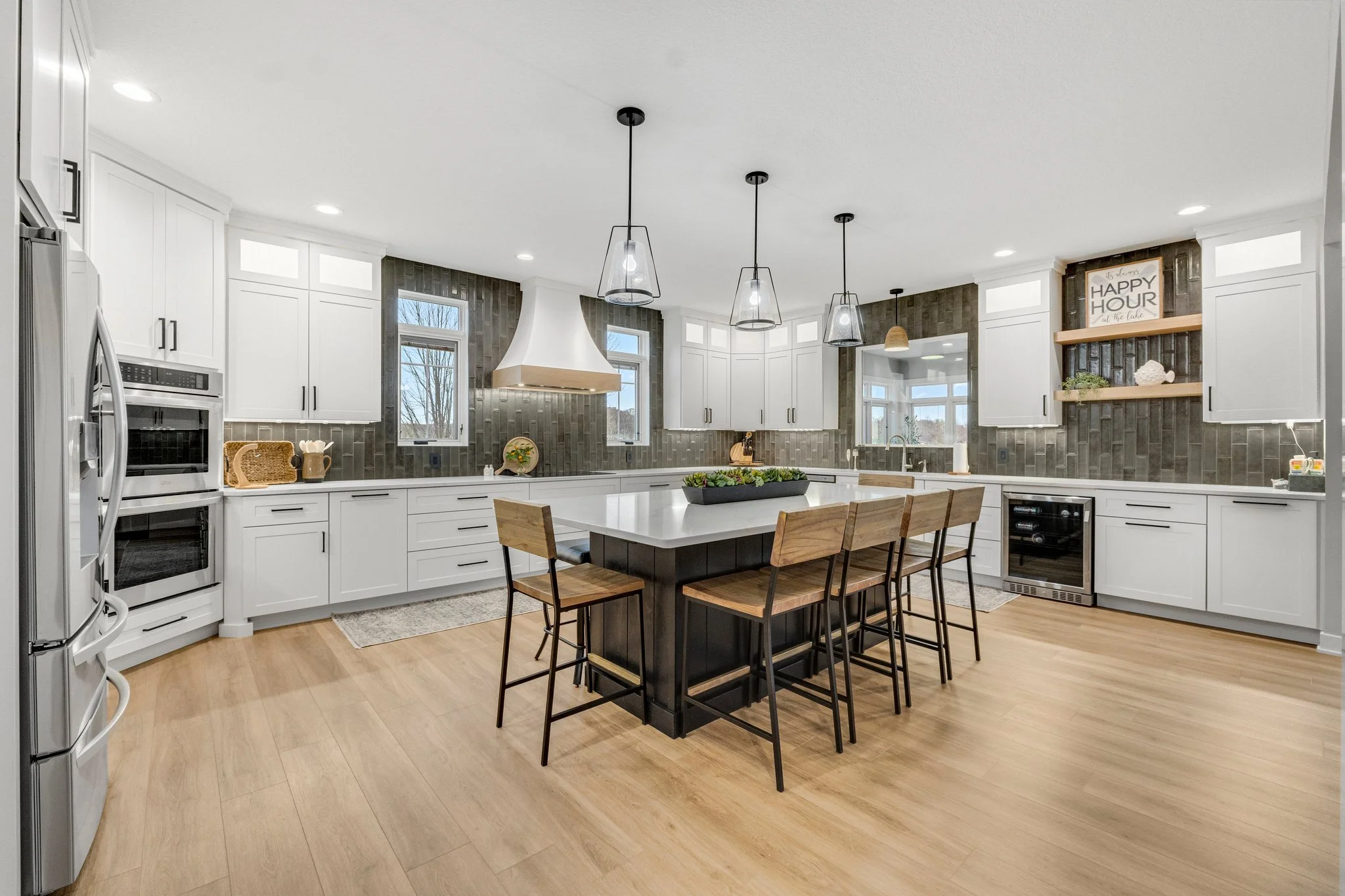 Modern kitchen with white cabinets, black countertops, and a central island with a black base. Four wooden chairs with metal legs are around the island. There are pendant lights hanging above the island. The kitchen has wood flooring and grey textured backsplash tiles. Two windows provide natural light, and there is a sign on the wall that reads 'Happy Hour.'