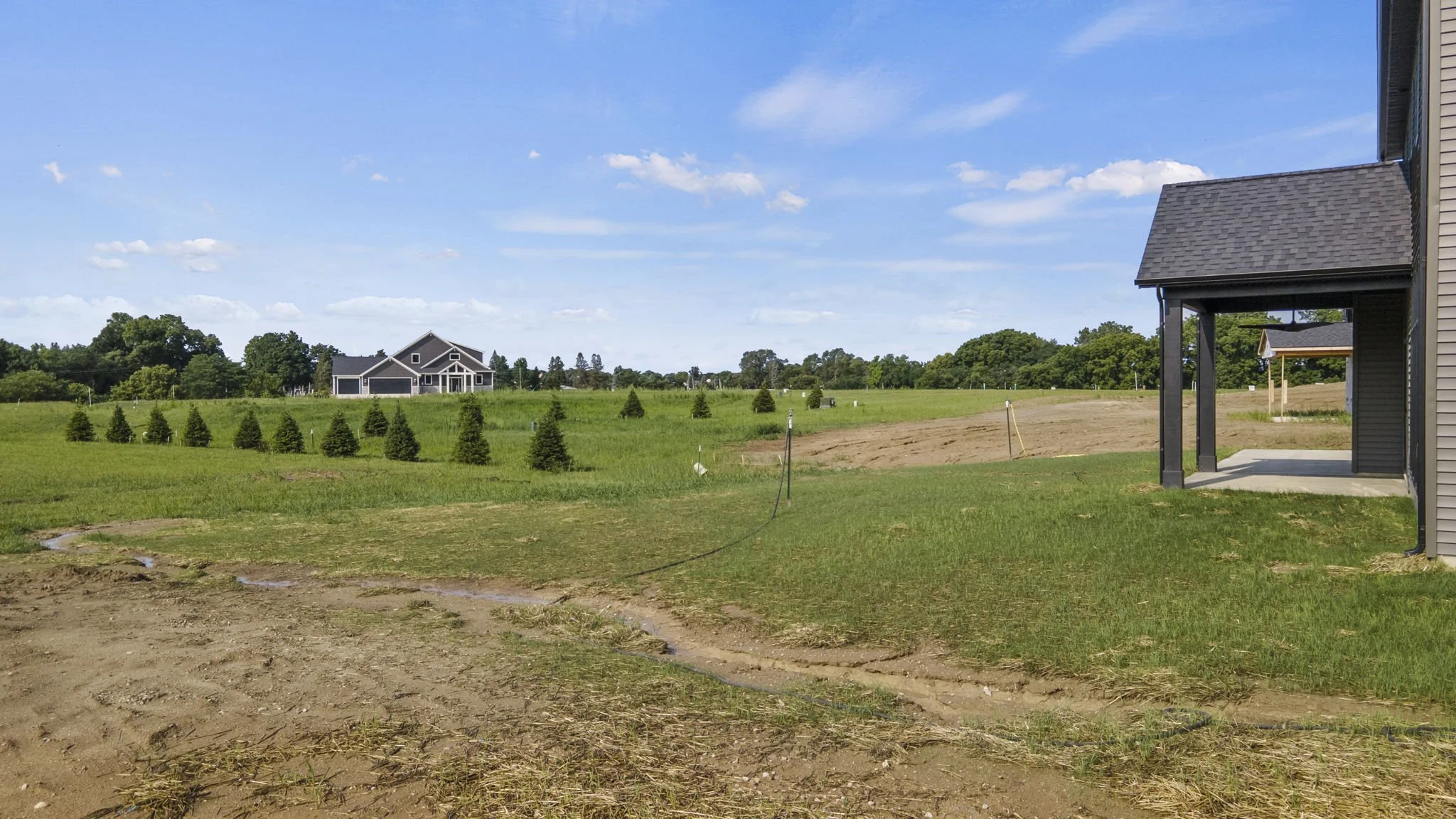 A partly developed yard with dirt and patches of grass, with a house on the right side and a line of small trees in the background, under a blue sky with clouds.