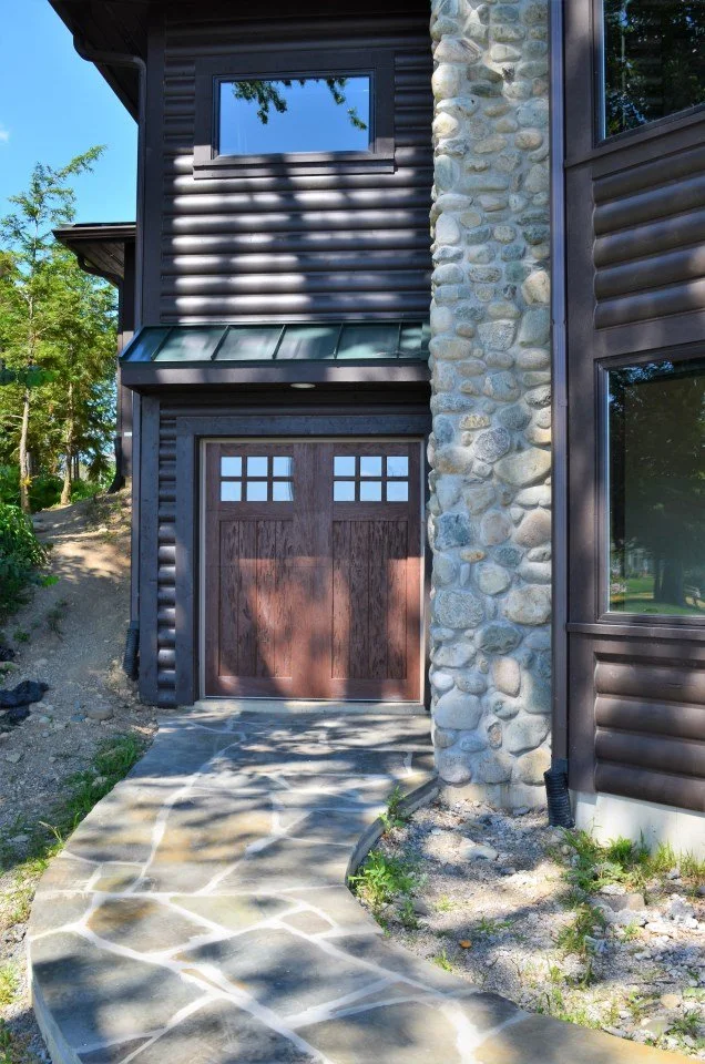 Front view of a house with a wooden garage door, stone chimney, and dark siding, with a concrete walkway leading to the garage door.