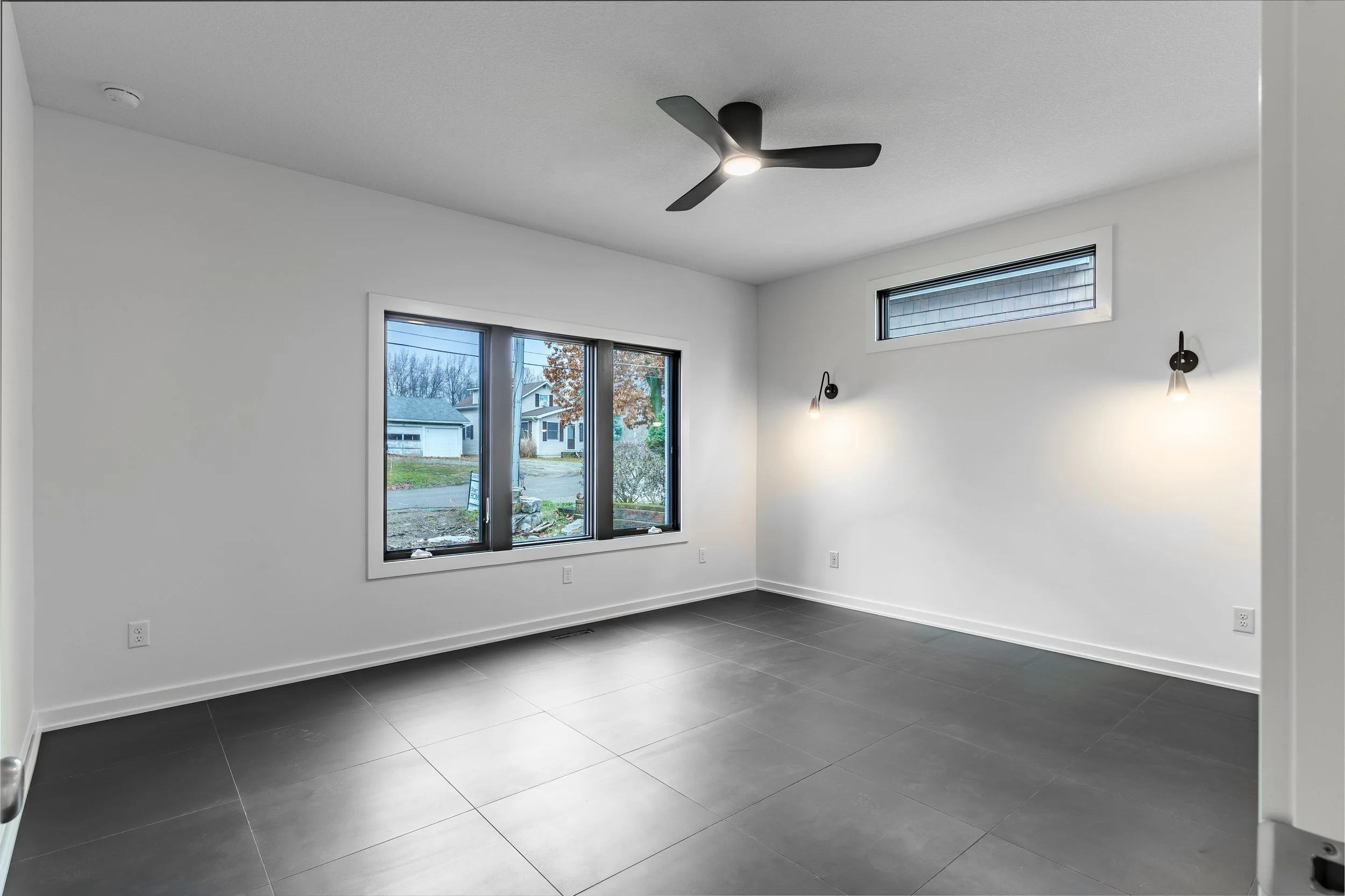 Empty modern room with large window, white walls, grey tiled floor, ceiling fan, and wall-mounted lights.
