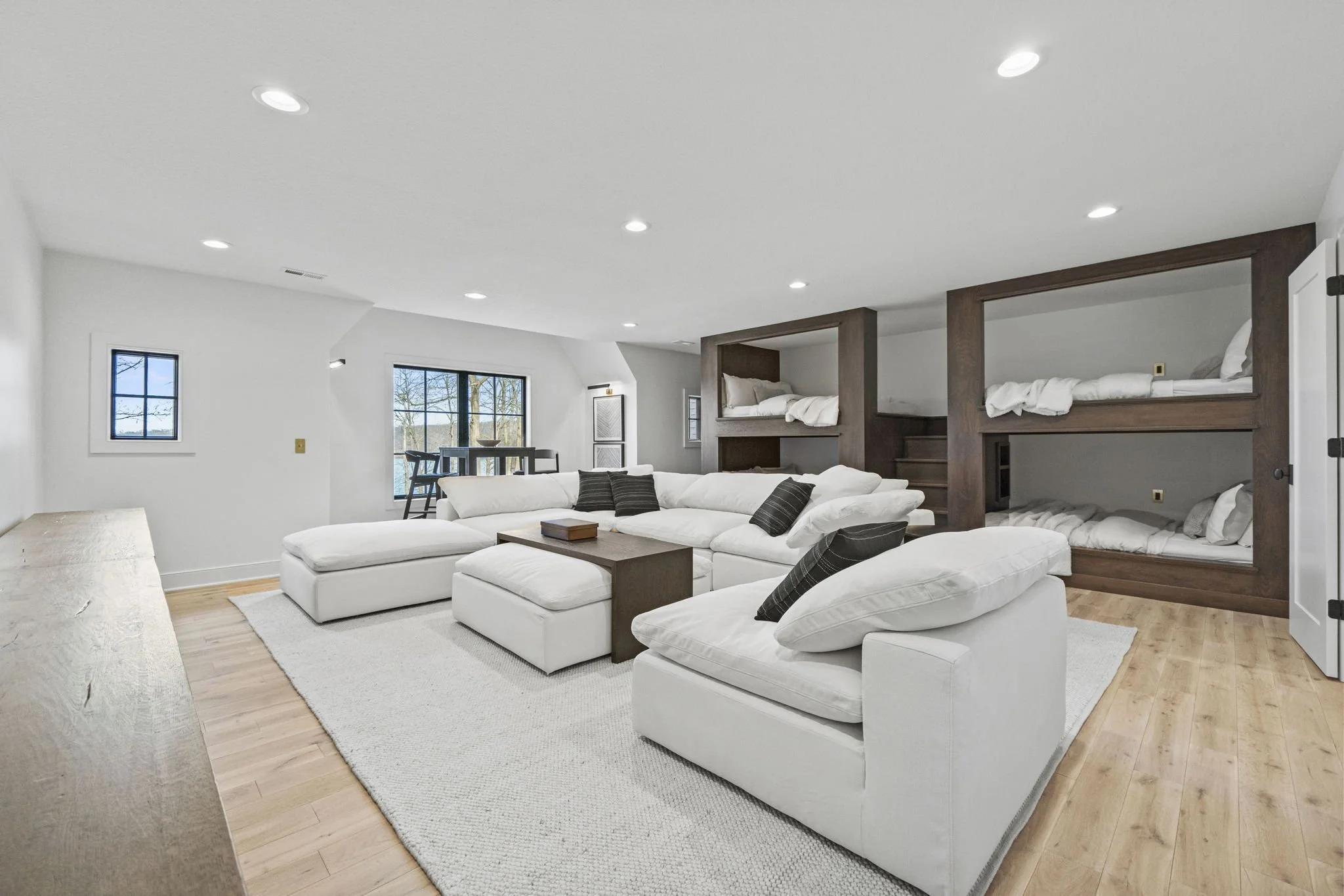 Living room with white sectional sofa, wooden coffee table, and built-in bunk beds with white bedding, light wood flooring, and windows showing outdoor trees.
