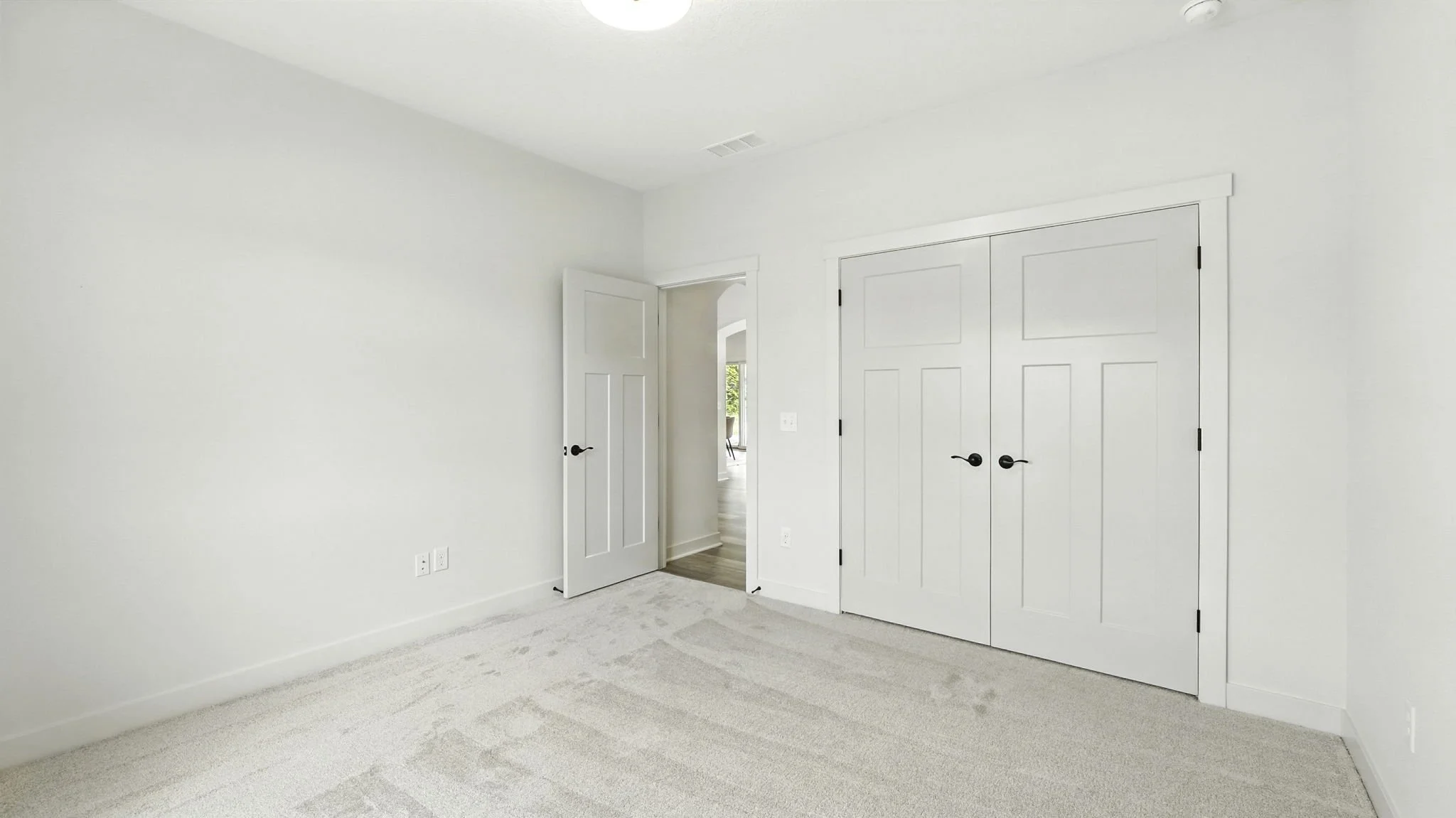 Empty white bedroom with closet doors and an open door leading to a hallway with a glimpse of another room.