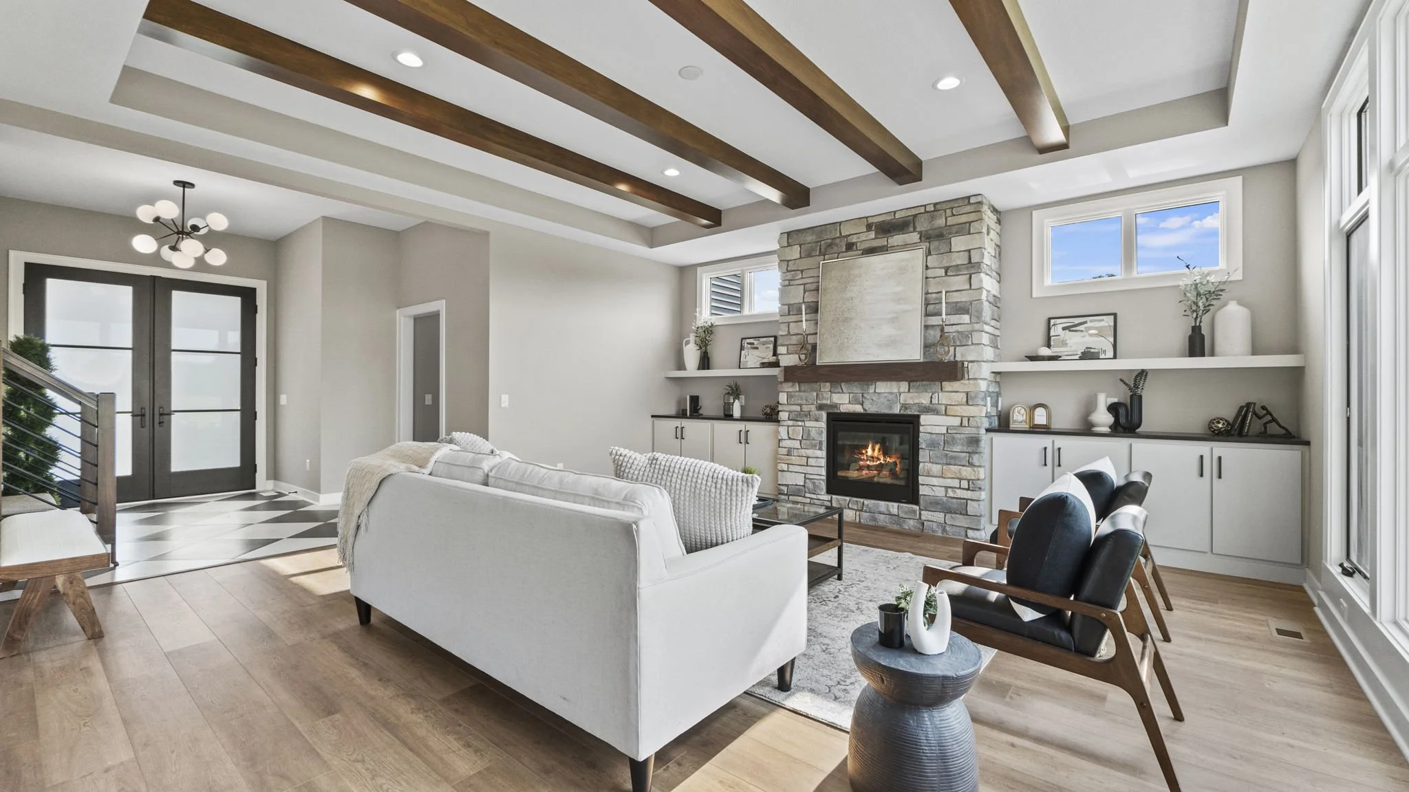 A modern living room with a stone fireplace, white sofa, black armchairs, built-in shelves, hardwood floors, and large windows with a view of the sky.