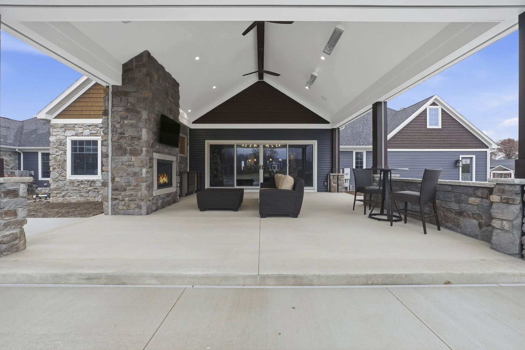 Covered outdoor patio with stone fireplace, black wicker chairs, and a bar table with chairs, overlooking neighboring houses with blue siding.