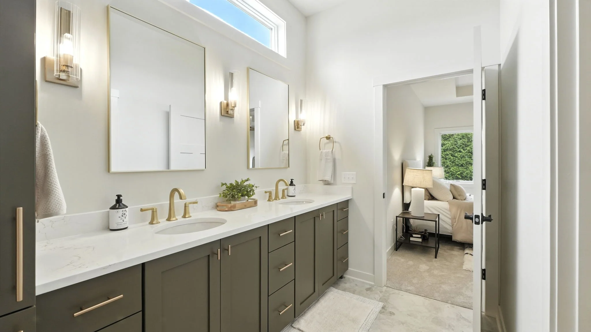 Modern bathroom with double vanity, white marble countertop, gold fixtures, large mirrors, and wall-mounted lights. A small plant and hand soap are on the counter, and a towel hanging on a gold ring