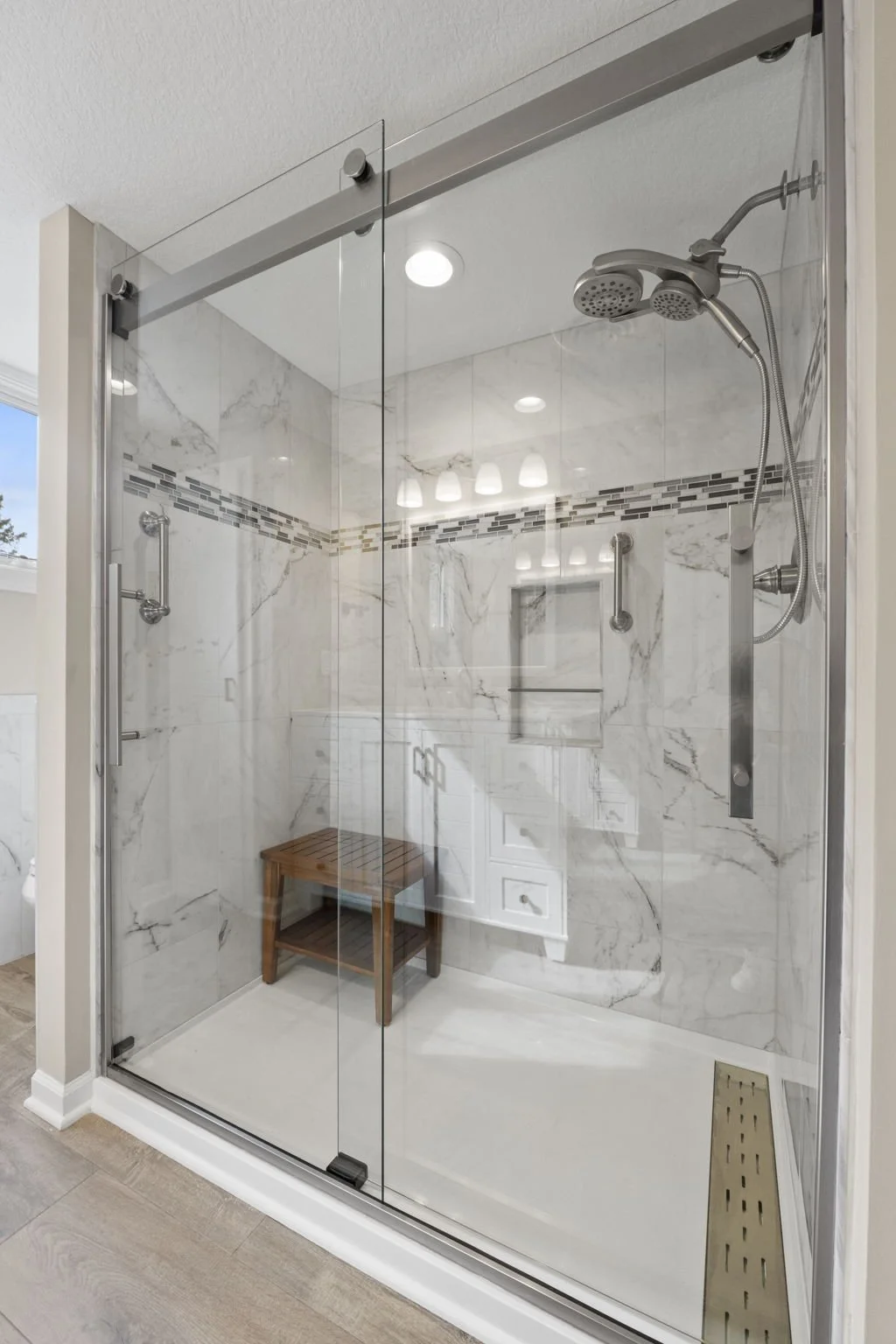 Glass shower enclosure with marble walls and a built-in bench, featuring a handheld showerhead, a sliding door, and adjustable grab bars.