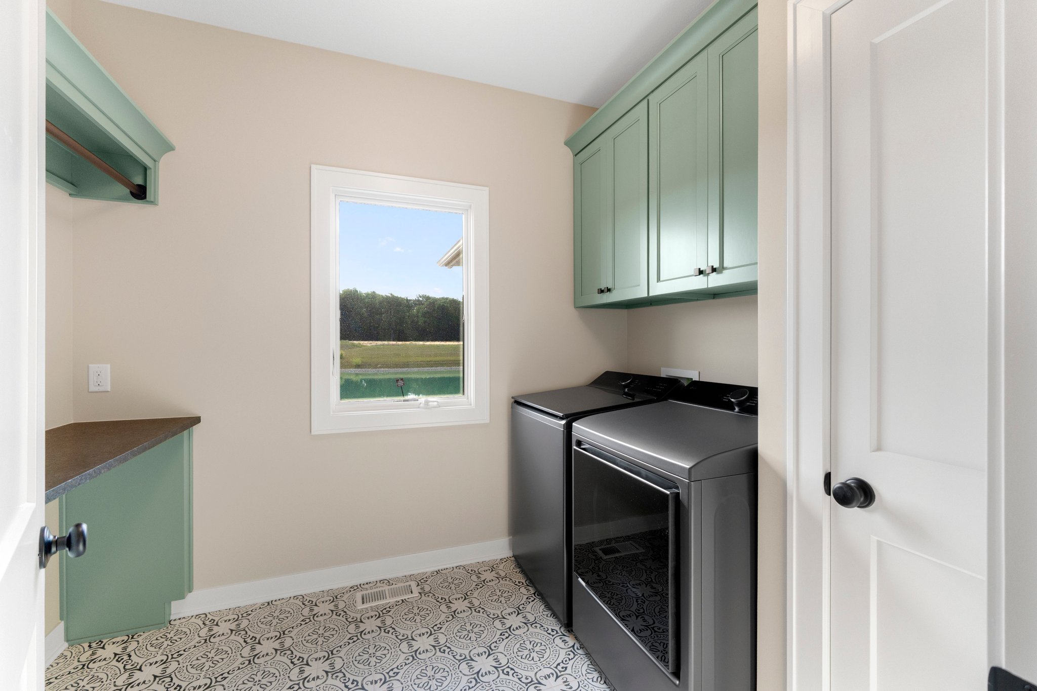 Laundry room with green cabinets, black washer and dryer, patterned floor, beige walls, small window showing outdoor landscape