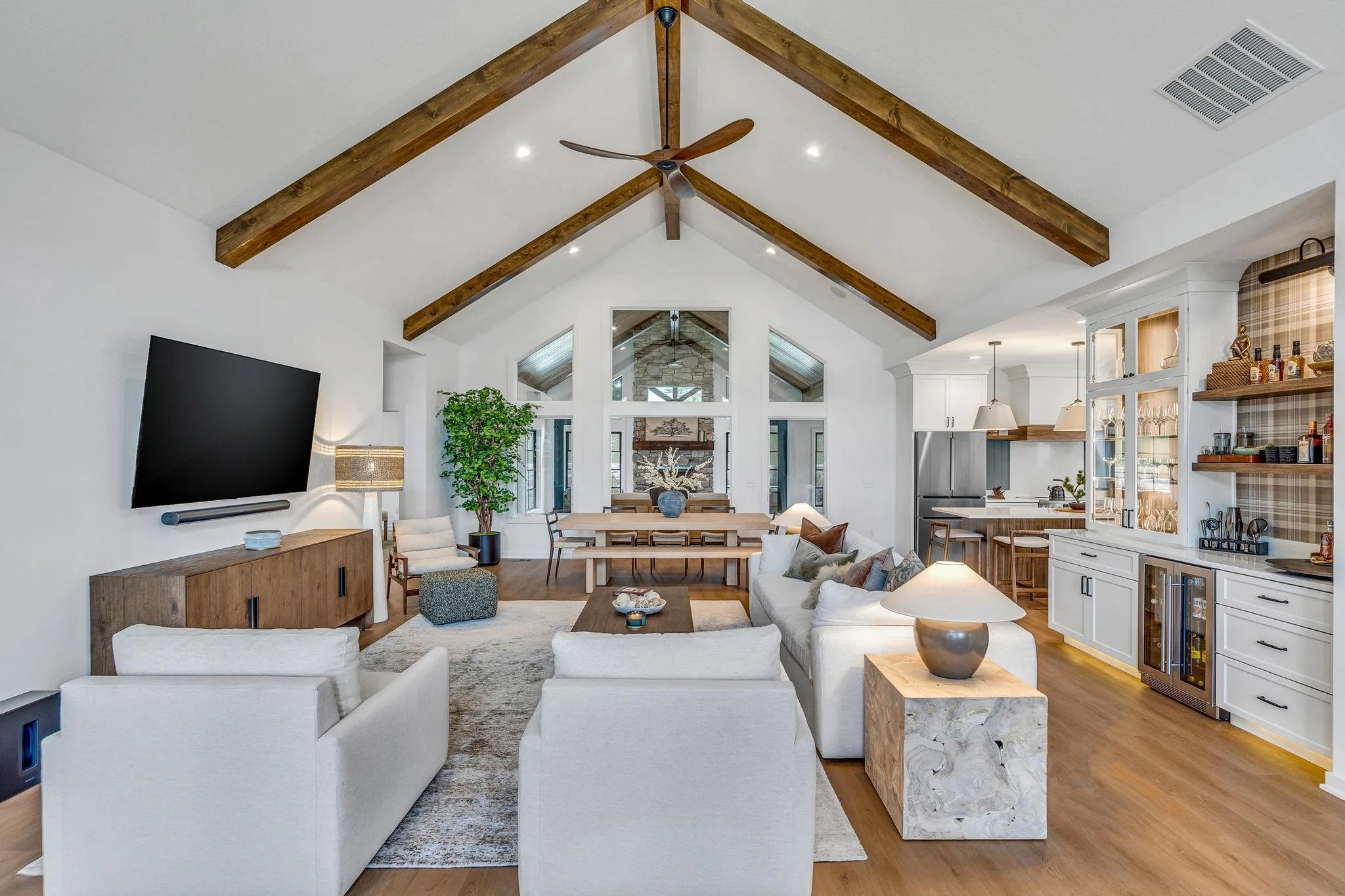 A spacious living room with white walls, wooden vaulted ceiling with exposed beams, and hardwood flooring. The room features a large flat-screen TV on the left, a white sofa set, a coffee table, a side table with a lamp, and a potted plant. In the ba