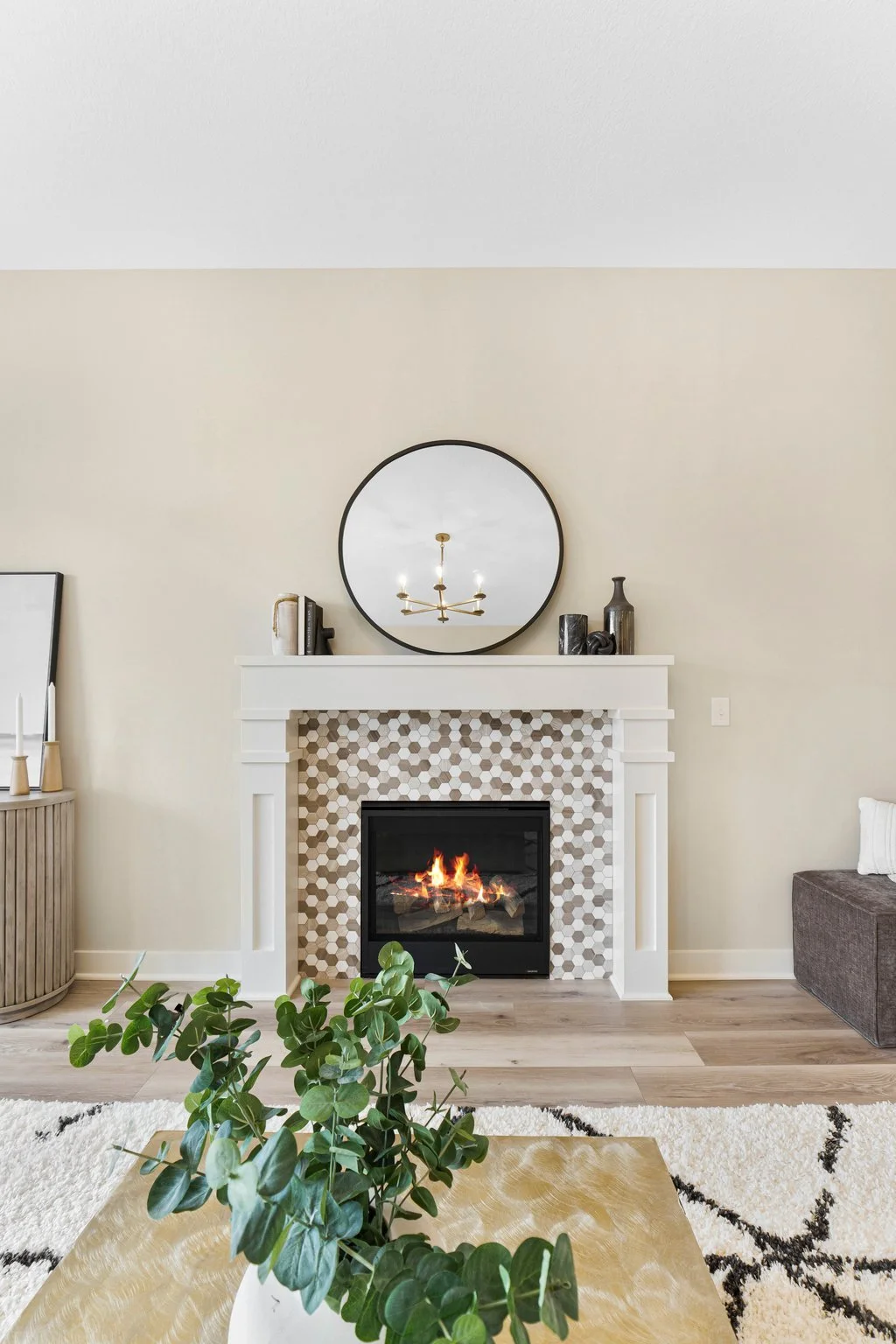 Living room with a white fireplace, round mirror, decorative vases, and a small fireplace with flames, a beige wall, wooden flooring, plants, and furniture.