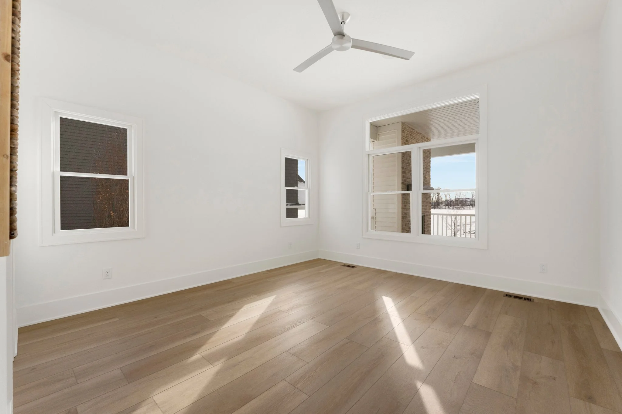 Empty room with white walls, light wood flooring, two small windows, and a ceiling fan.