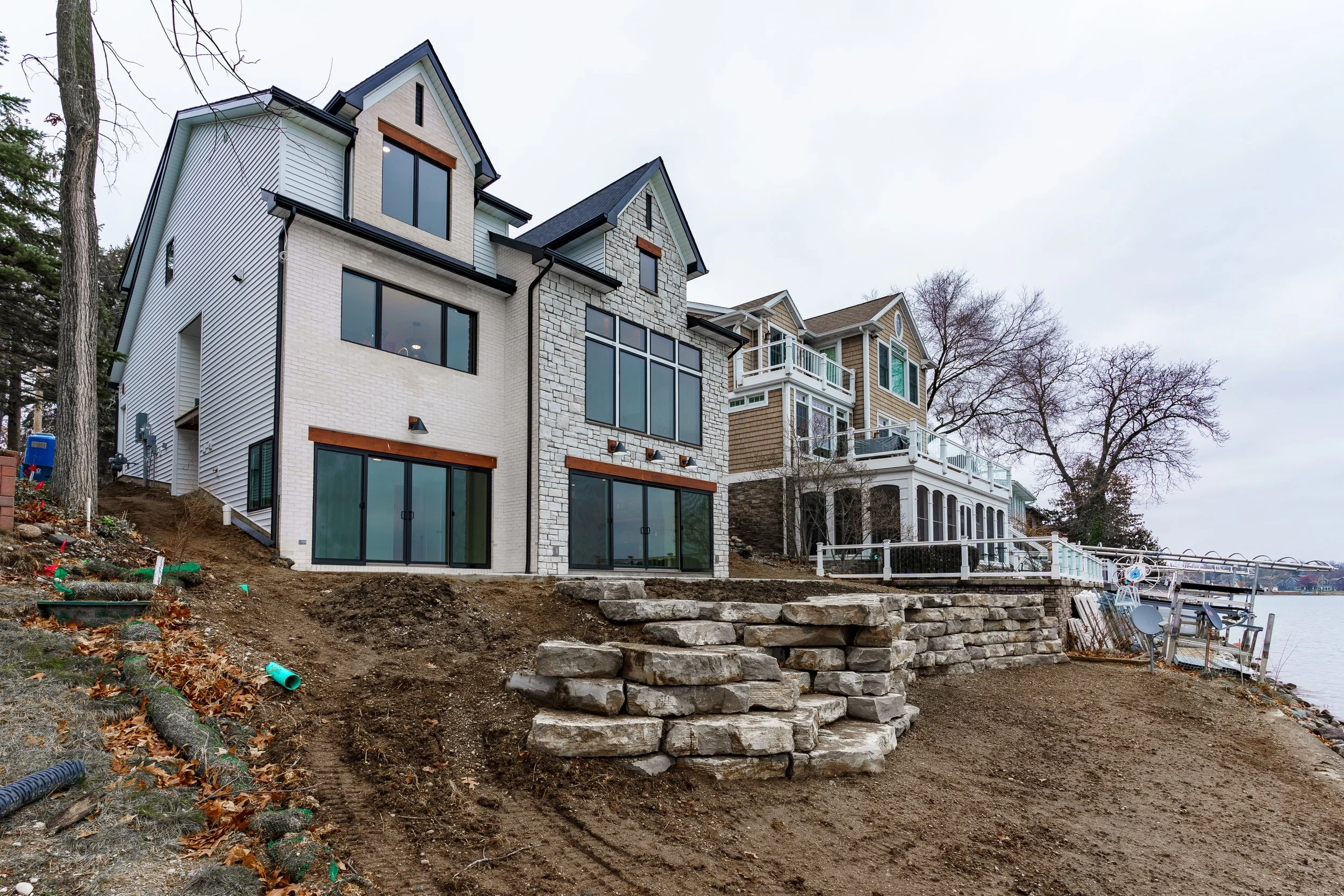 Newly constructed multi-story house with large windows, located on a hillside next to a body of water, with an unfinished yard and stone retaining wall in the foreground.