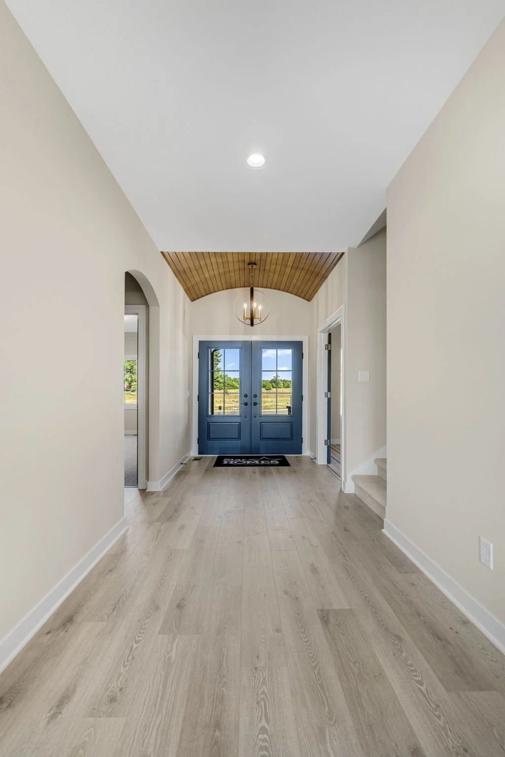 Entrance hallway of a house with light hardwood flooring, beige walls, blue front door with glass panes, wooden arch ceiling, modern light fixture, and a view of outdoor fields through the door windows.