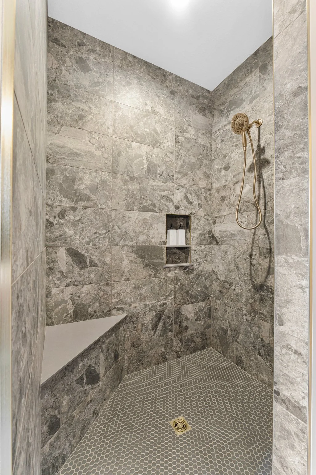A shower with gray marble tiles on the walls, a built-in corner shelf with two bottles, a gold showerhead, and a gold drain on a small gray hexagonal tile floor.