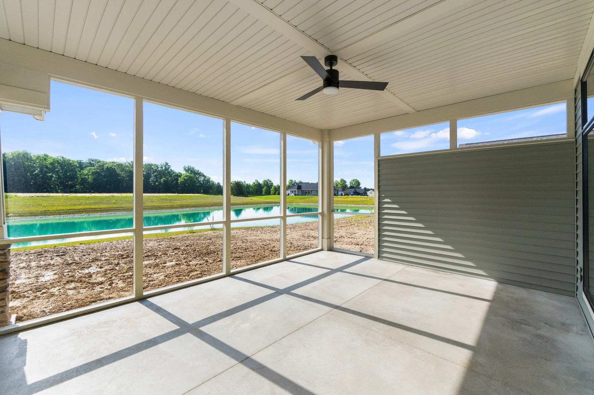 Empty screened porch with ceiling fan, overlooking a lake with green trees and houses in the distance.
