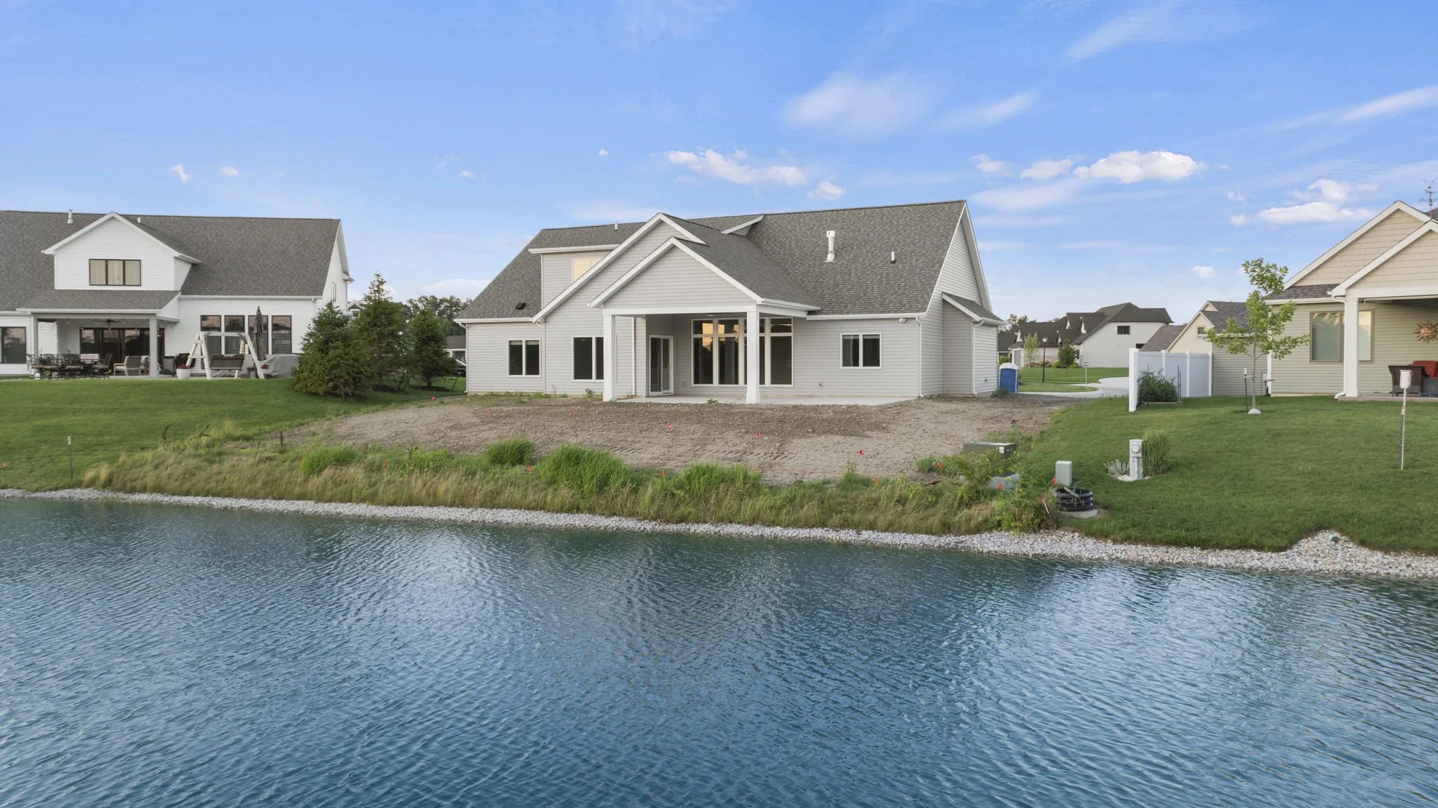 Newly constructed house with white siding and gray roof, situated by a body of water, with neighboring homes in a suburban setting and a partially landscaped yard.