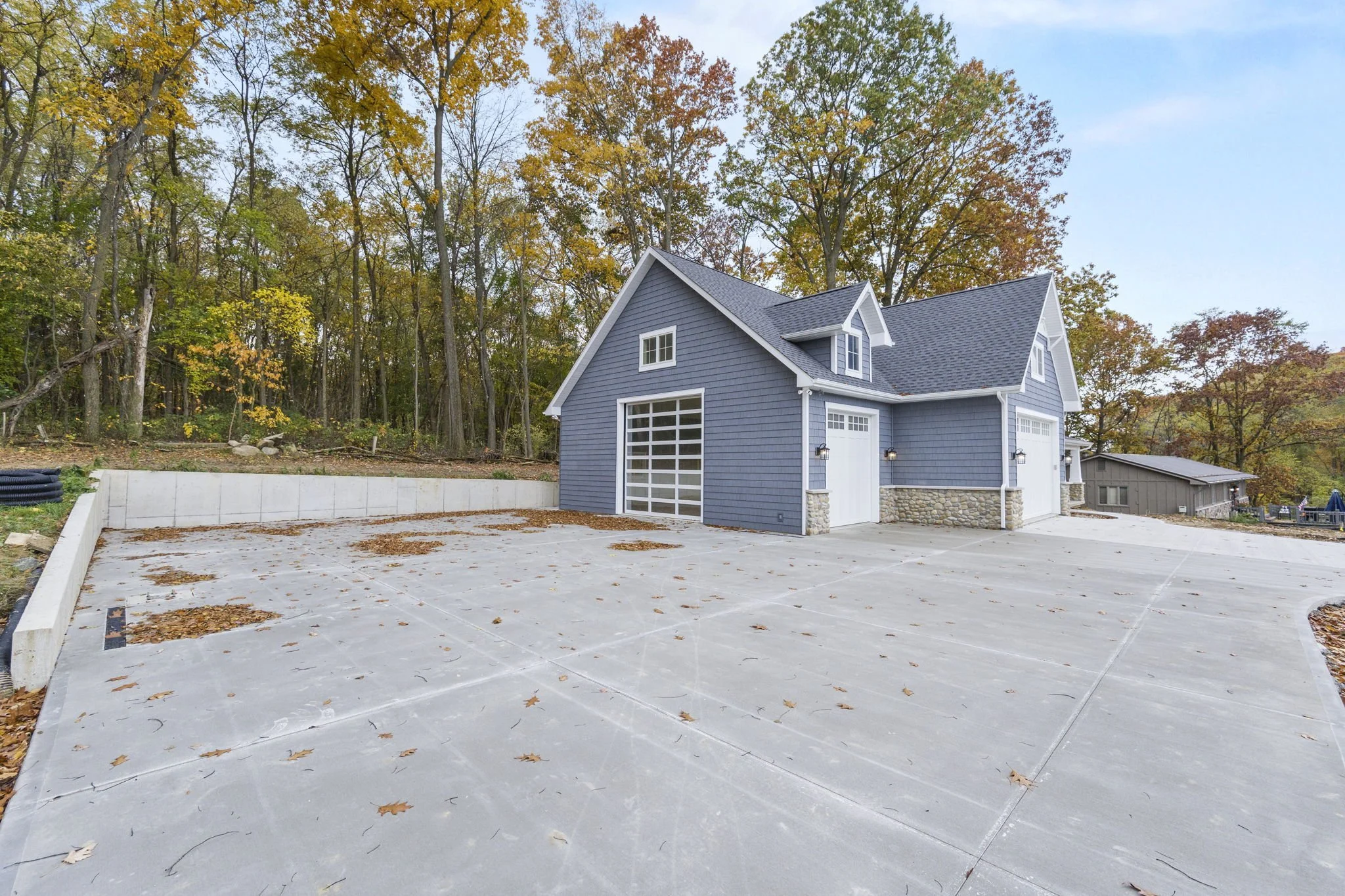 A newly built blue garage with white trim, stone accents, and a concrete driveway surrounded by fall trees with colorful leaves.