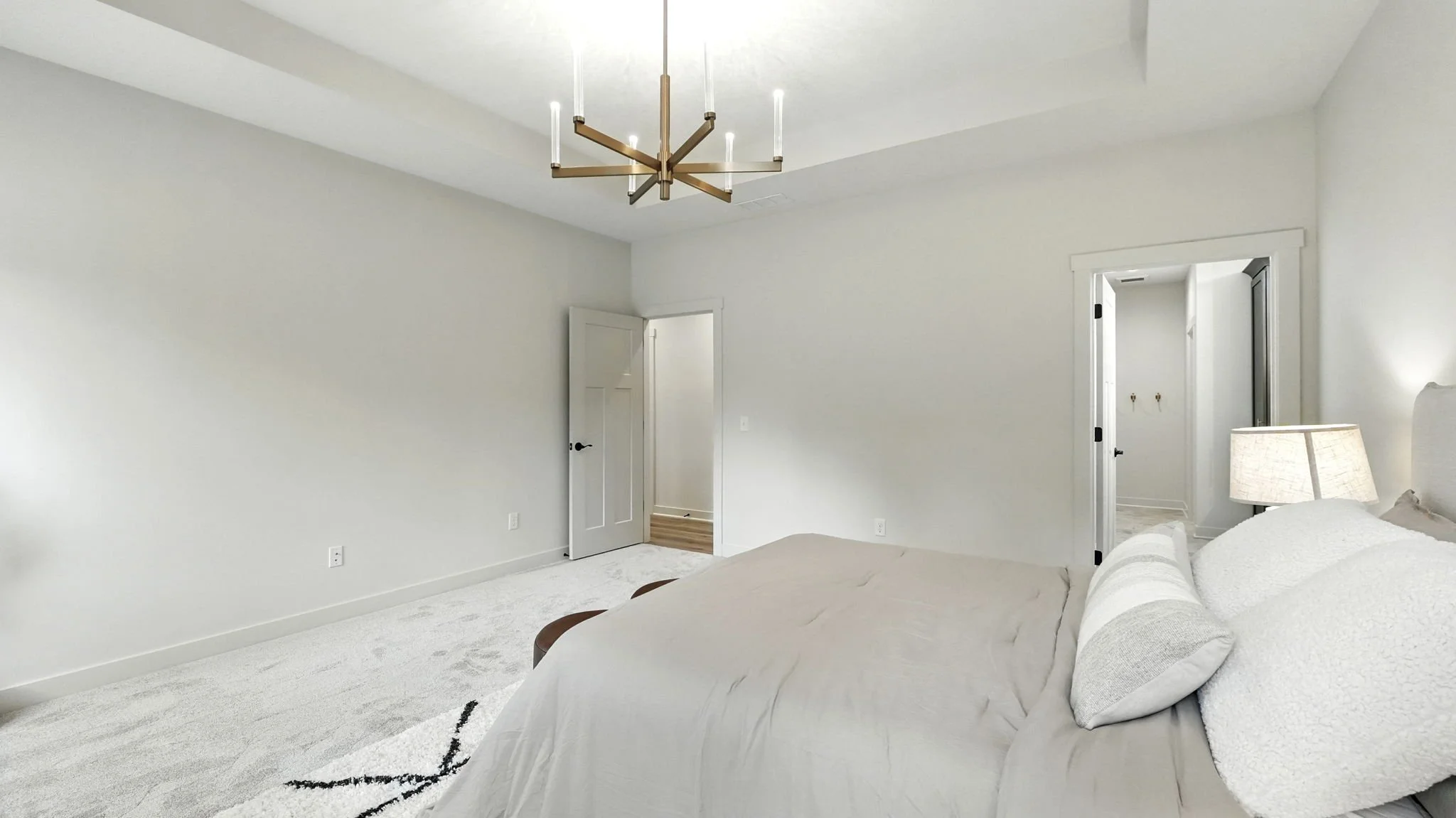 A minimalist bedroom with white walls, a bed with white bedding, a wall-mounted bedside lamp, a modern chandelier, and two doorways leading to other rooms.