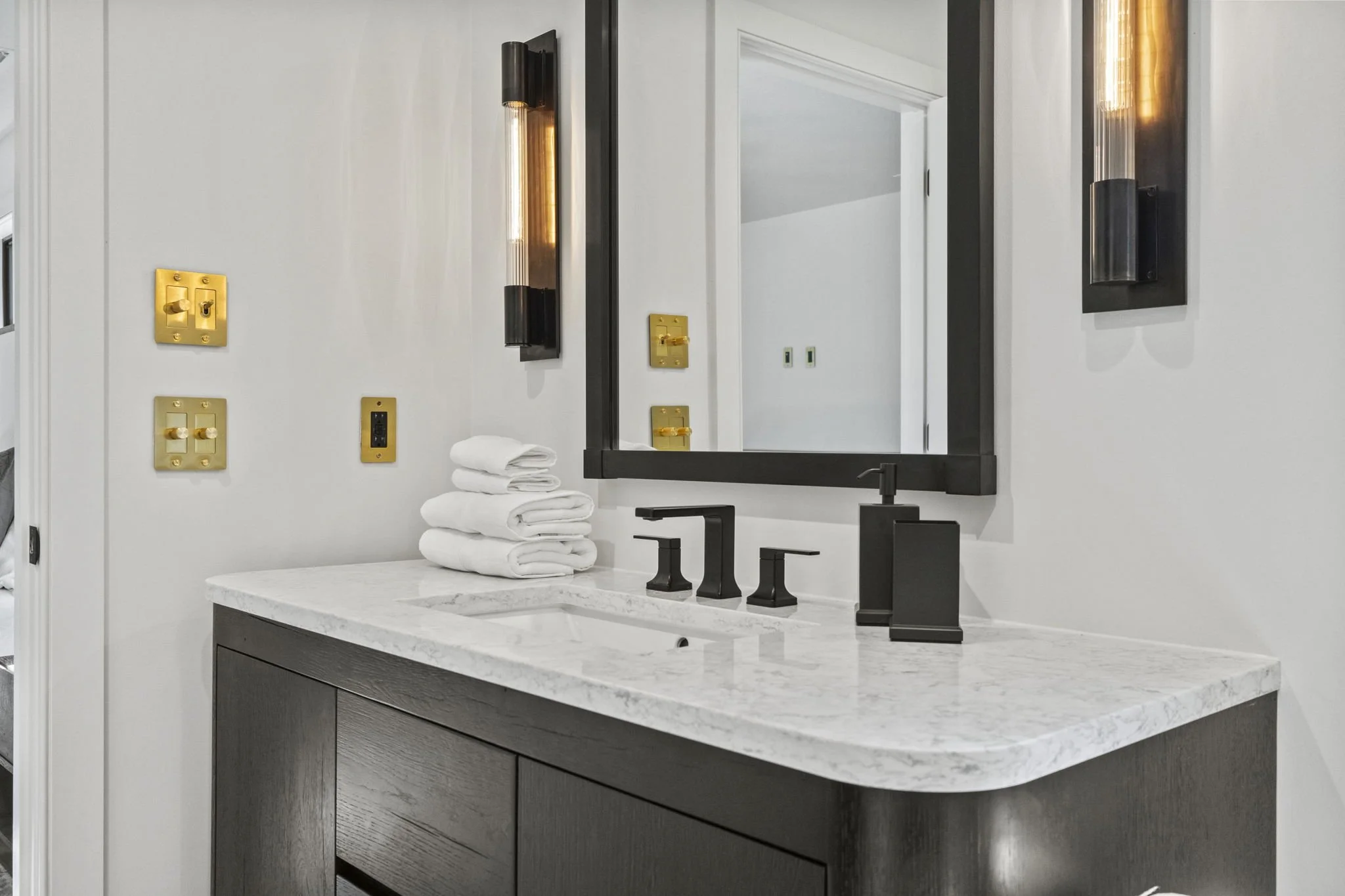 Bathroom vanity with white marble countertop, black fixtures, and a large black-framed mirror. Four white towels are folded on the counter, and wall-mounted black light fixtures flank the mirror. Gold-colored electrical switches and outlets are visib