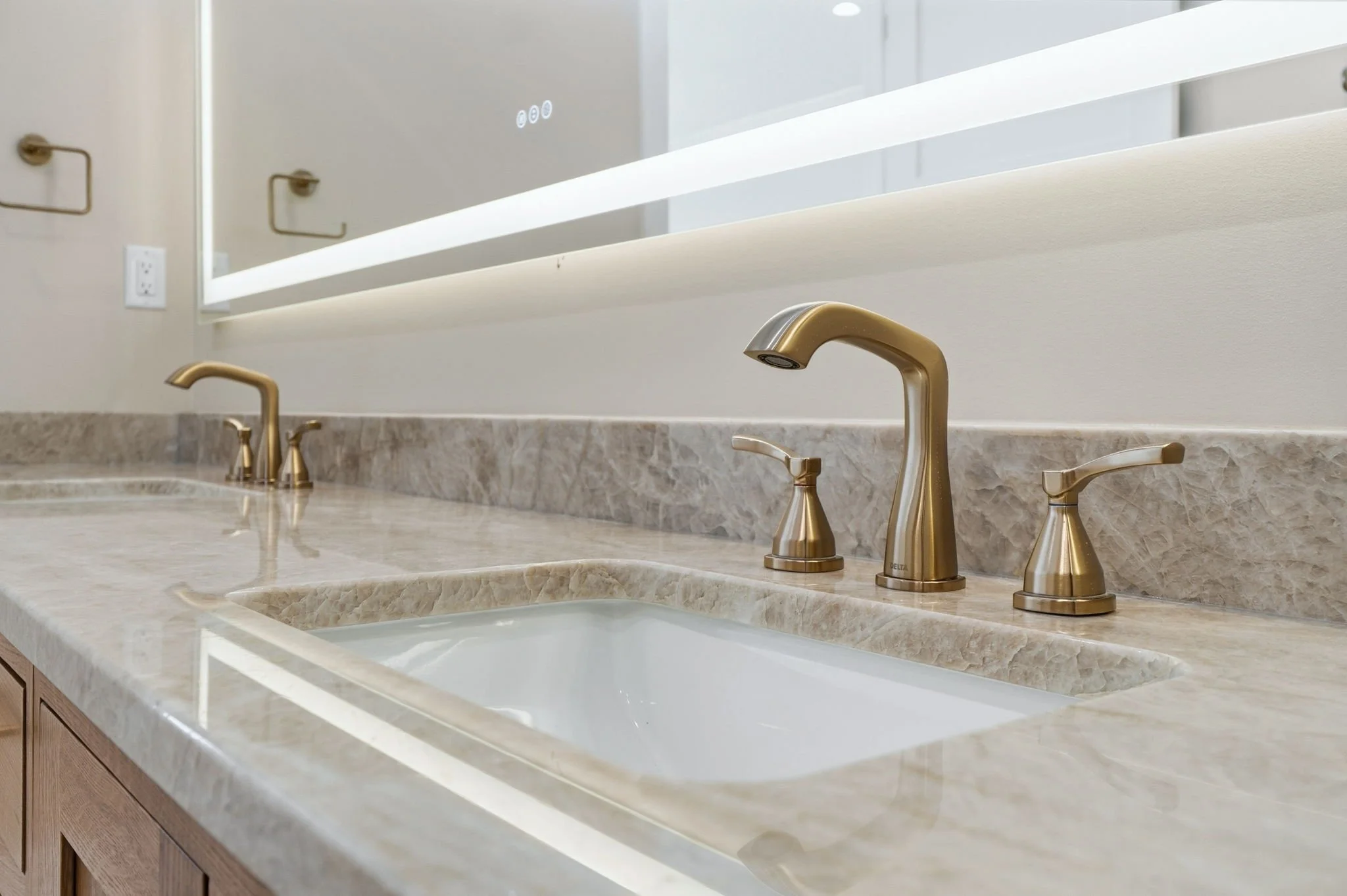 A bathroom countertop with two sinks, gold-colored faucets, and a mirror above with LED lighting.
